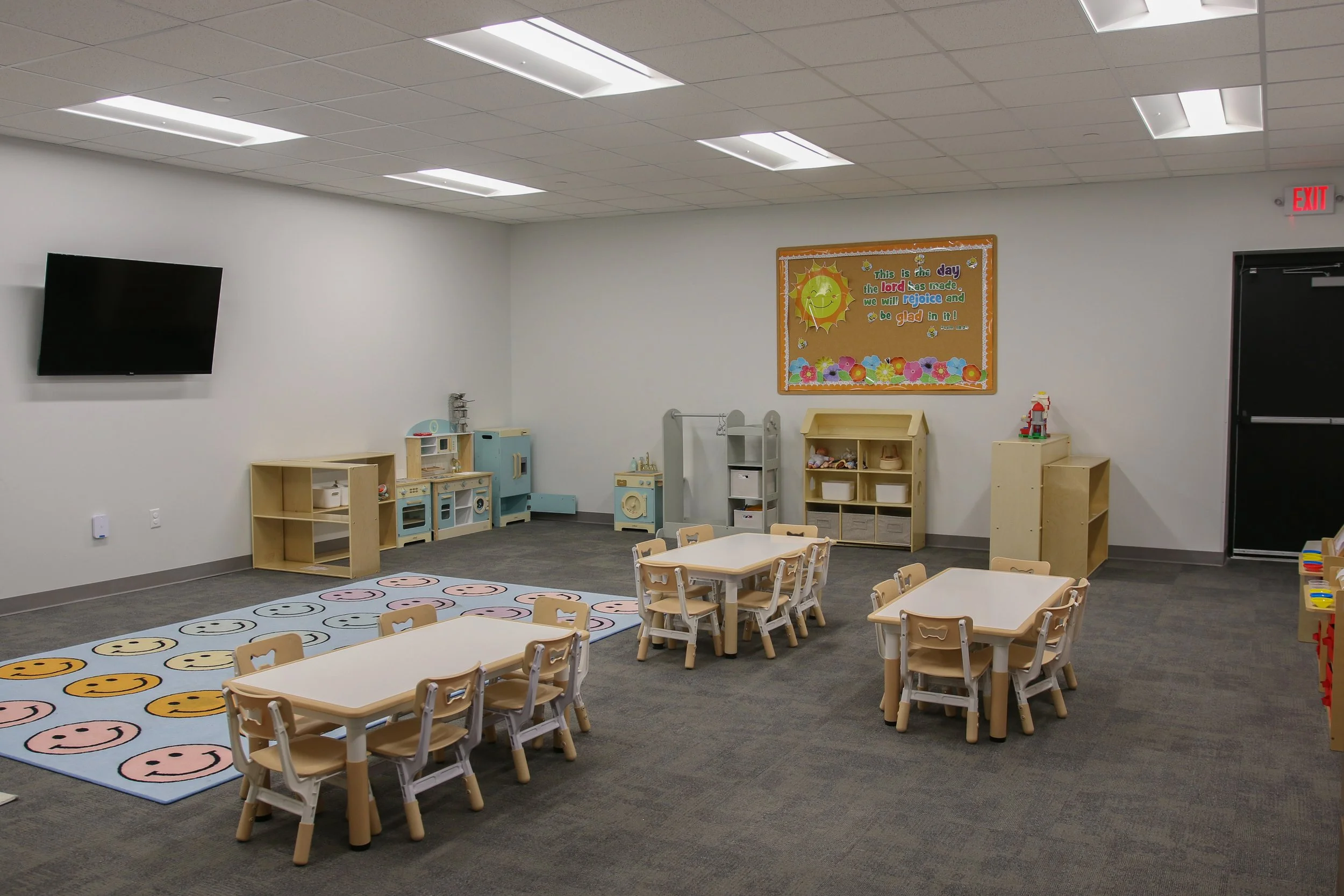 A classroom with small tables and chairs, a colorful smiley face rug, a bulletin board with a sun and flowers, toy kitchen set, and shelves, designed for young children.