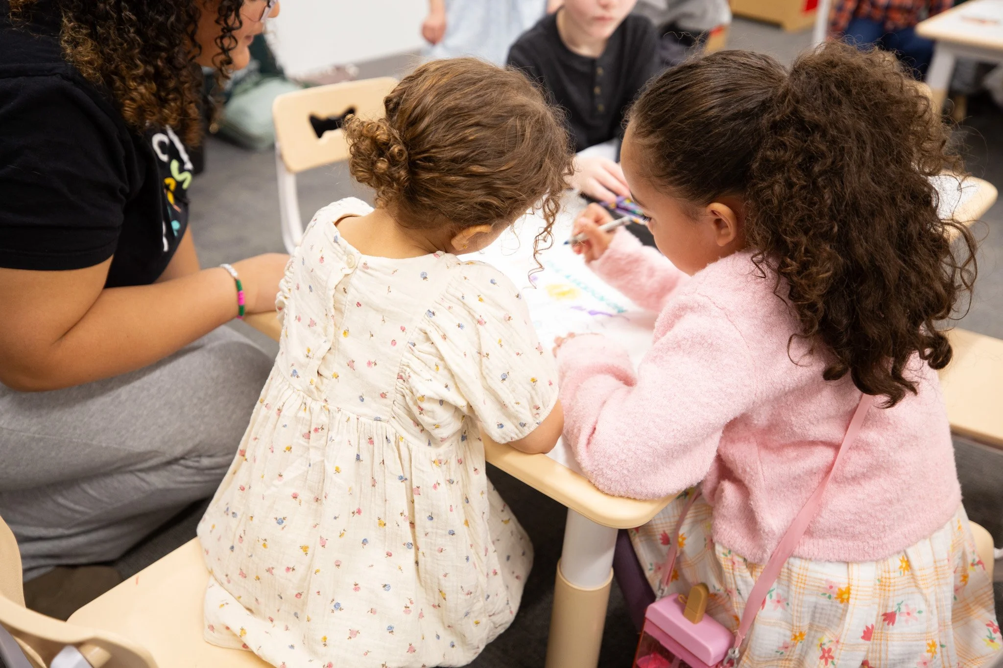 A group of young girls with curly hair gathered around a table, engaging in a craft activity, in a classroom setting.