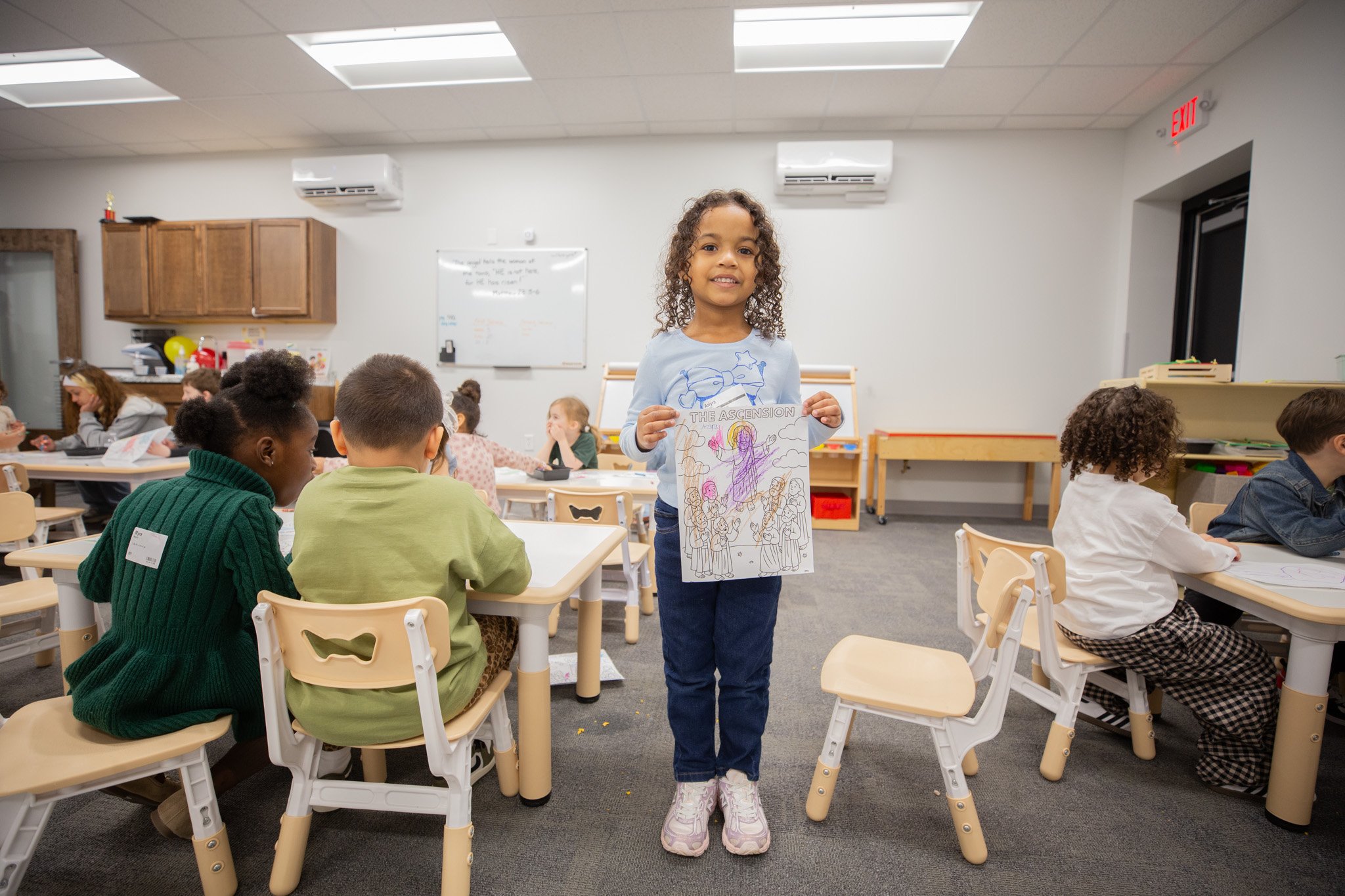 A young girl with curly hair standing in a classroom, holding a coloring page with the title 'The Ascension' and a drawing of Jesus with children, with other children sitting at desks around her.