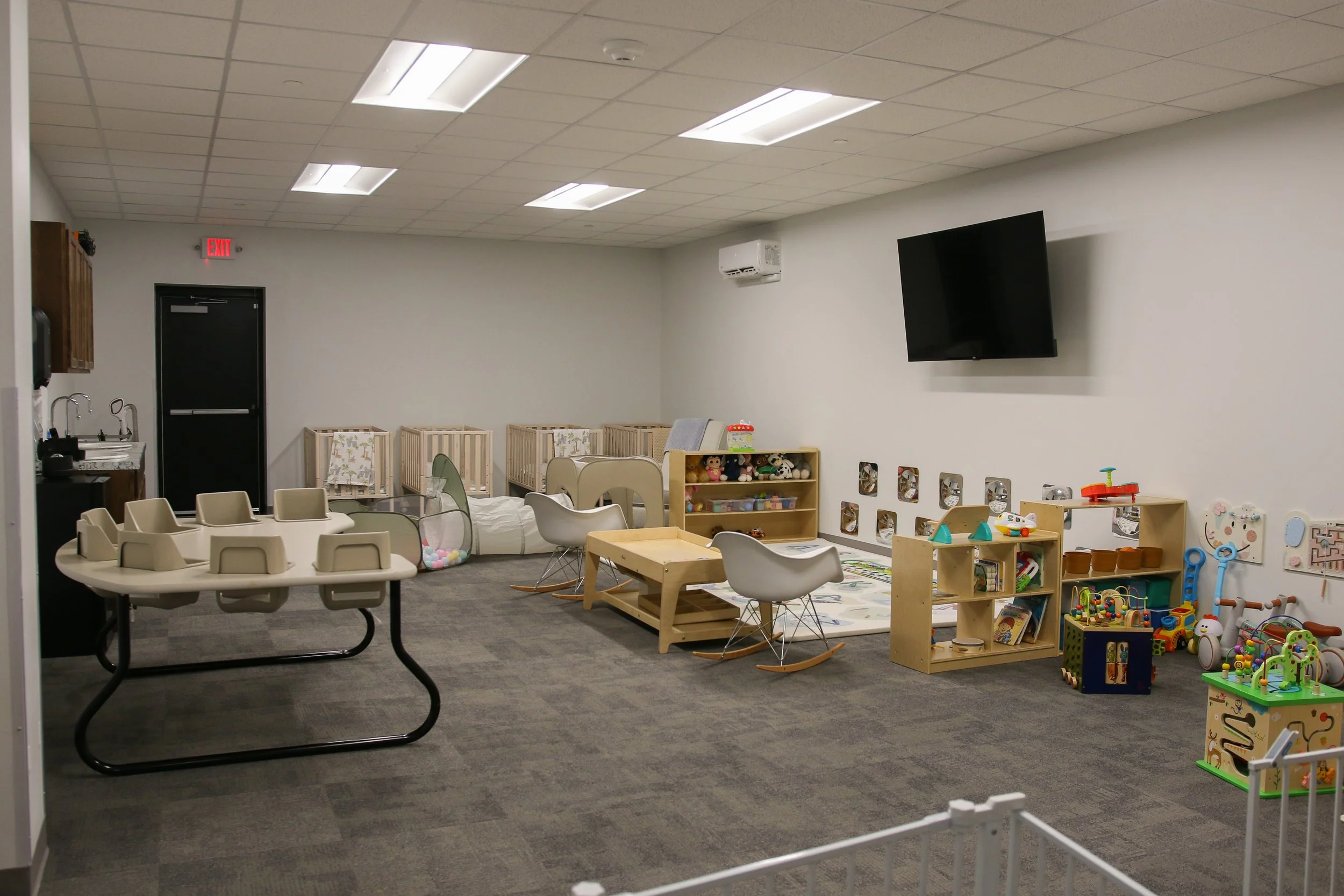 Indoor children's play and learning area with toys, tables, chairs, and small playhouses, featuring a wall-mounted TV and ceiling lights.