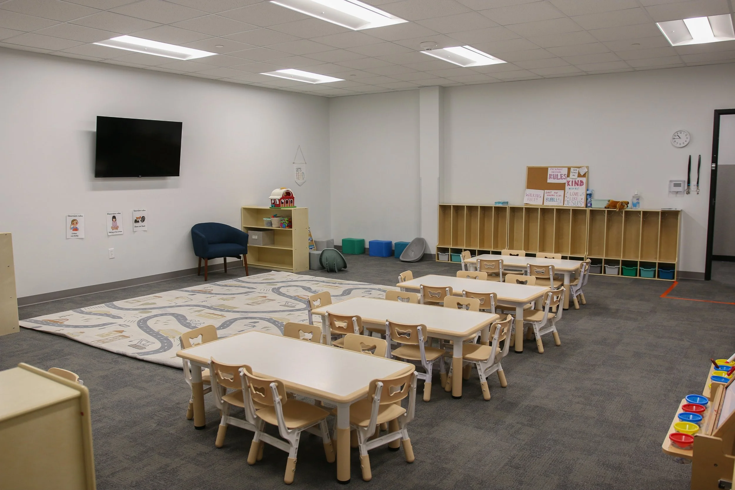 An empty classroom in a preschool or kindergarten with small tables and chairs, storage cubbies, a TV on the wall, and children's artwork and rules displayed on the walls.