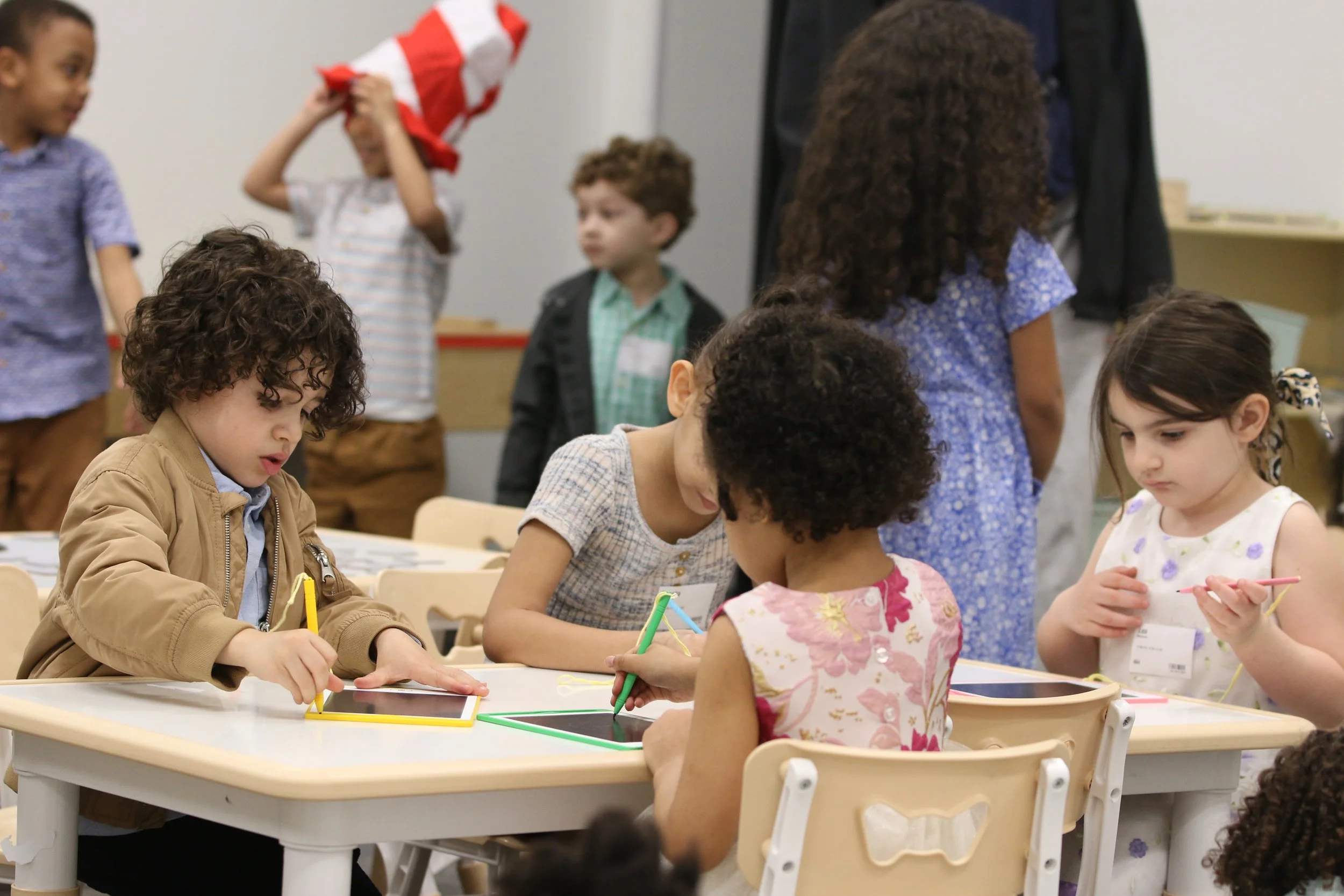 Group of young children at tables engaging in an activity, with some children drawing or writing on tablets, in a classroom setting.