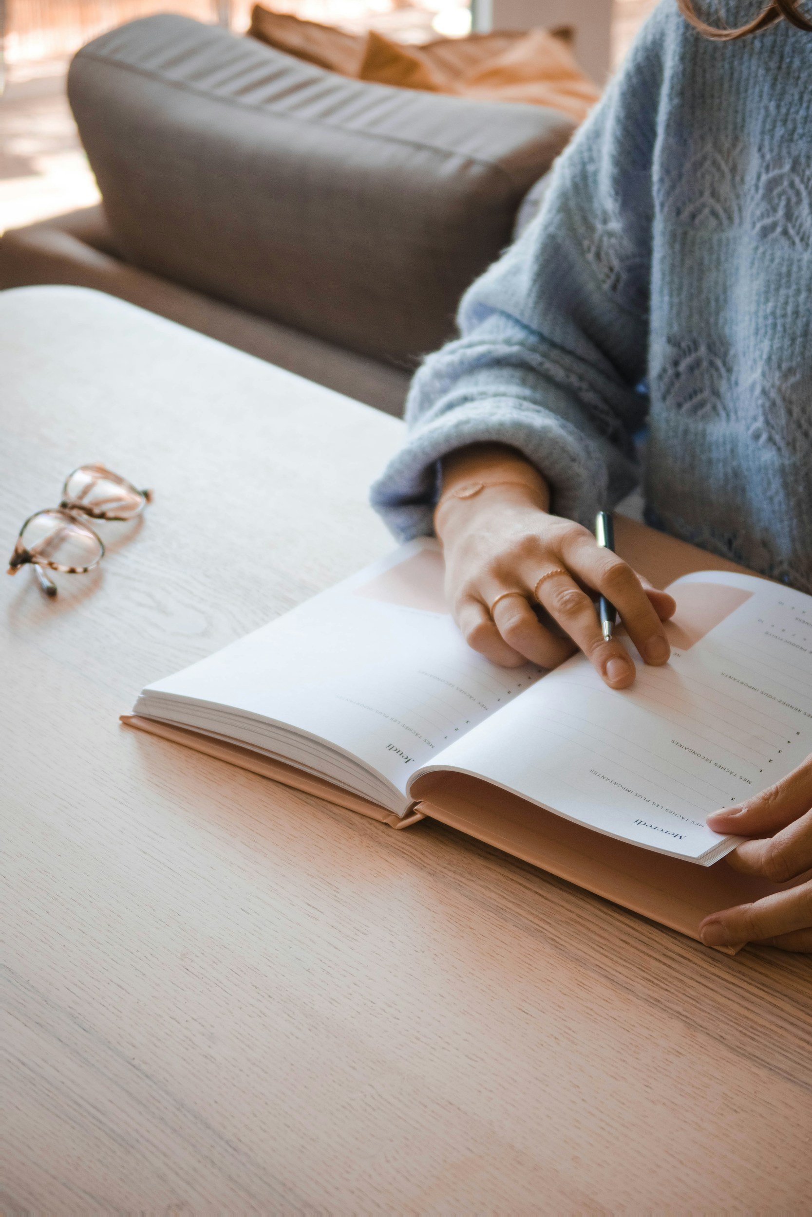 Person wearing a gray sweater writing in a workbook at a light-colored wooden table, with eyeglasses placed on the table. A sofa with beige cushions is visible in the background.