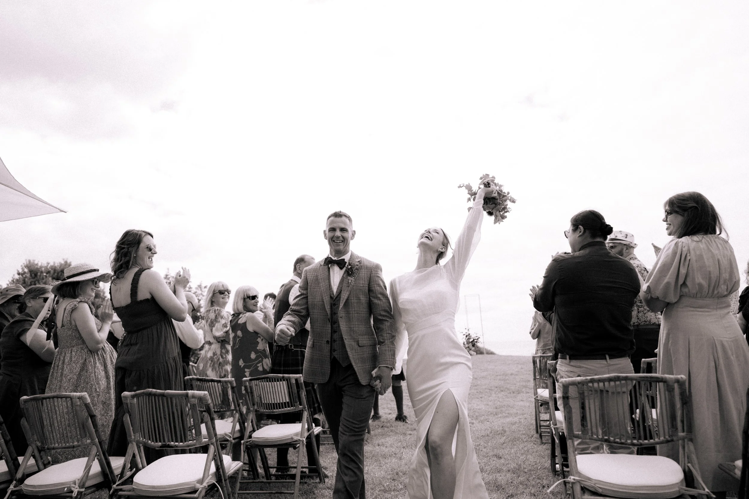 A bride and groom are celebrating at an outdoor wedding ceremony, with the bride holding a bouquet and raising her arm. Guests are smiling and clapping around them.
