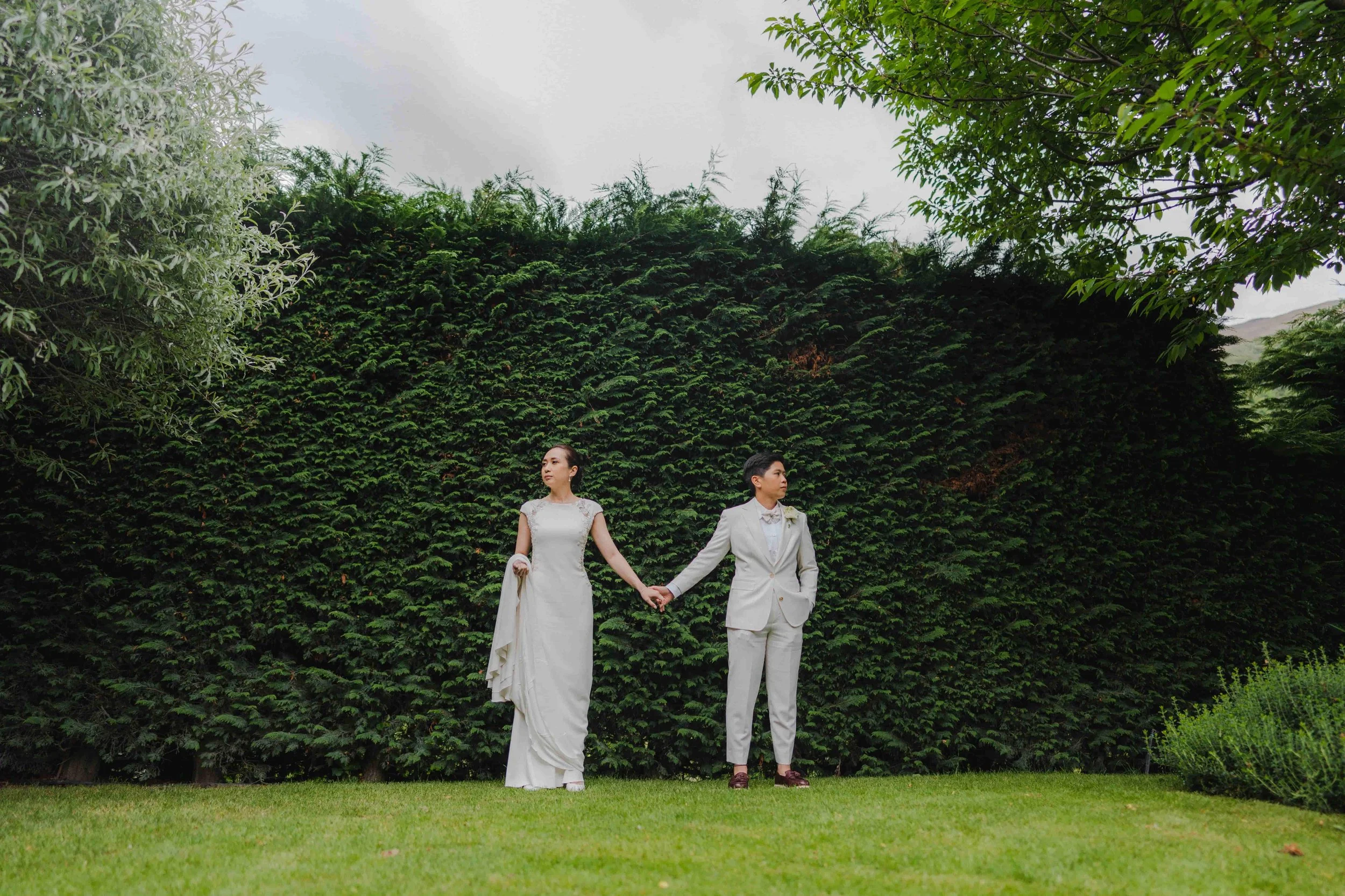 A bride and groom holding hands and standing outdoors on a grassy area, with a dense green hedge and trees behind them, overcast sky.