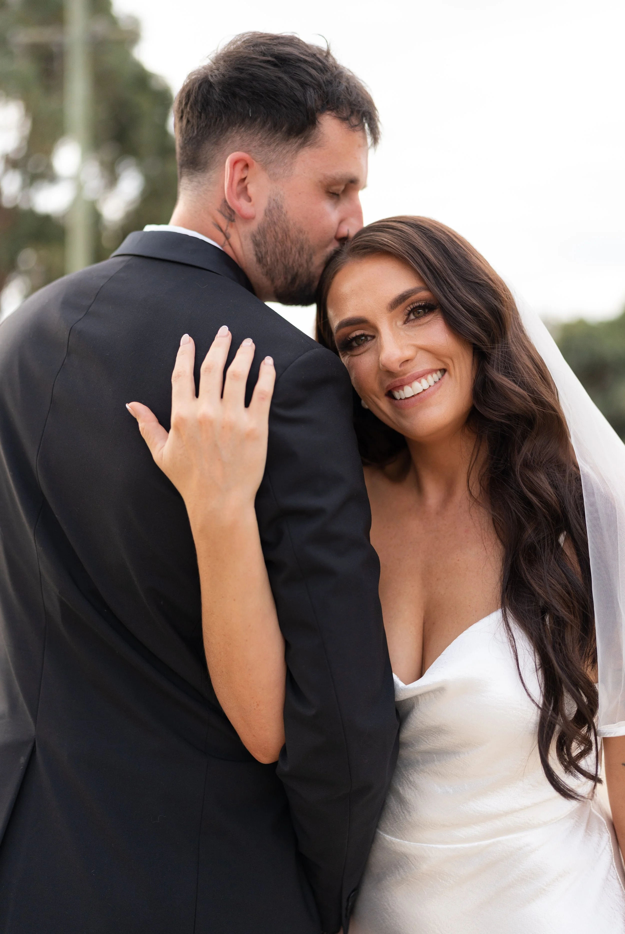 A happy bride and groom share a tender moment outdoors; the groom is kissing the bride's forehead, and she is smiling with her hand on his shoulder.