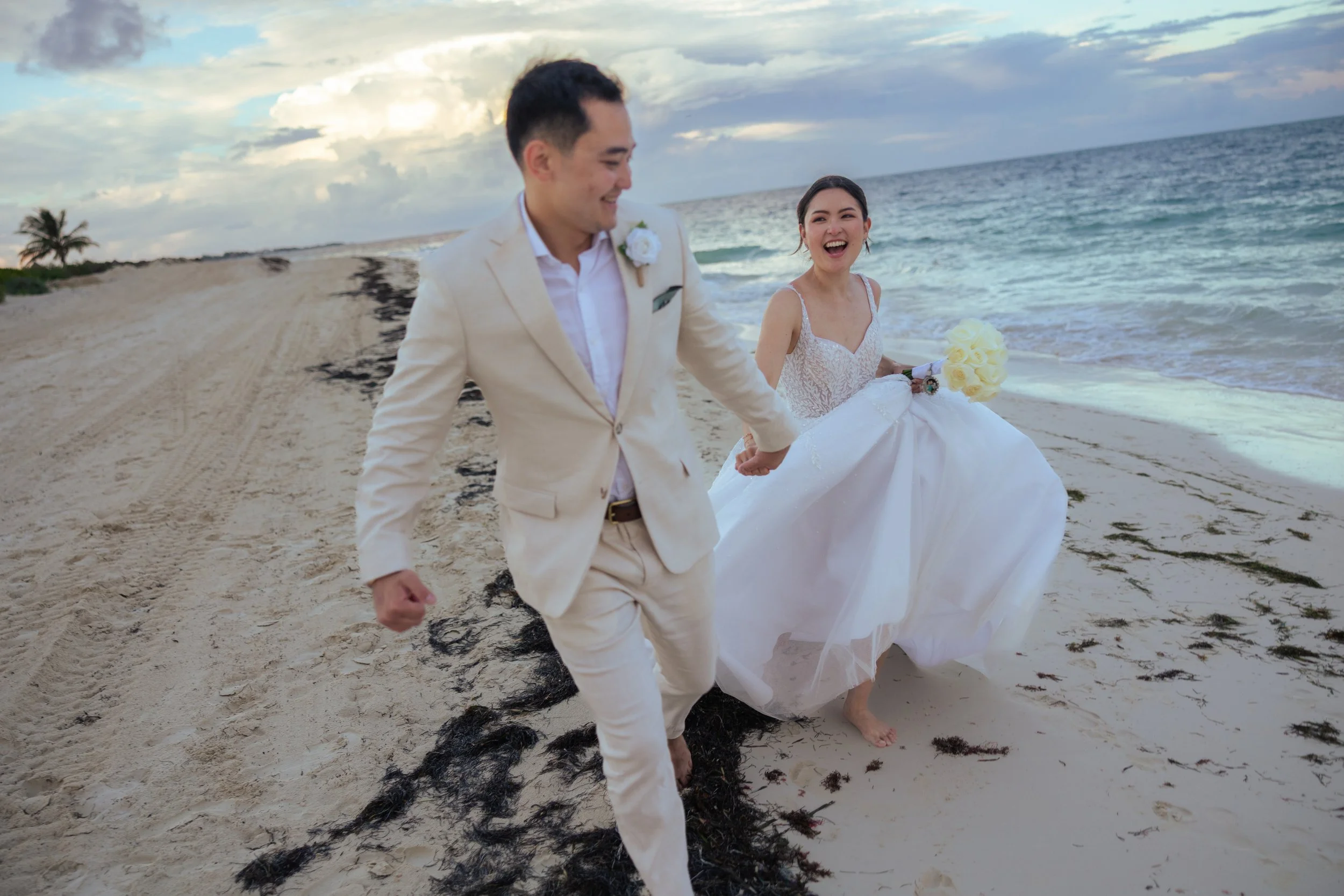 A joyous bride and groom in wedding attire walking hand in hand along a beach at sunset, smiling and holding a bouquet of white roses, with ocean waves and cloudy sky in the background.