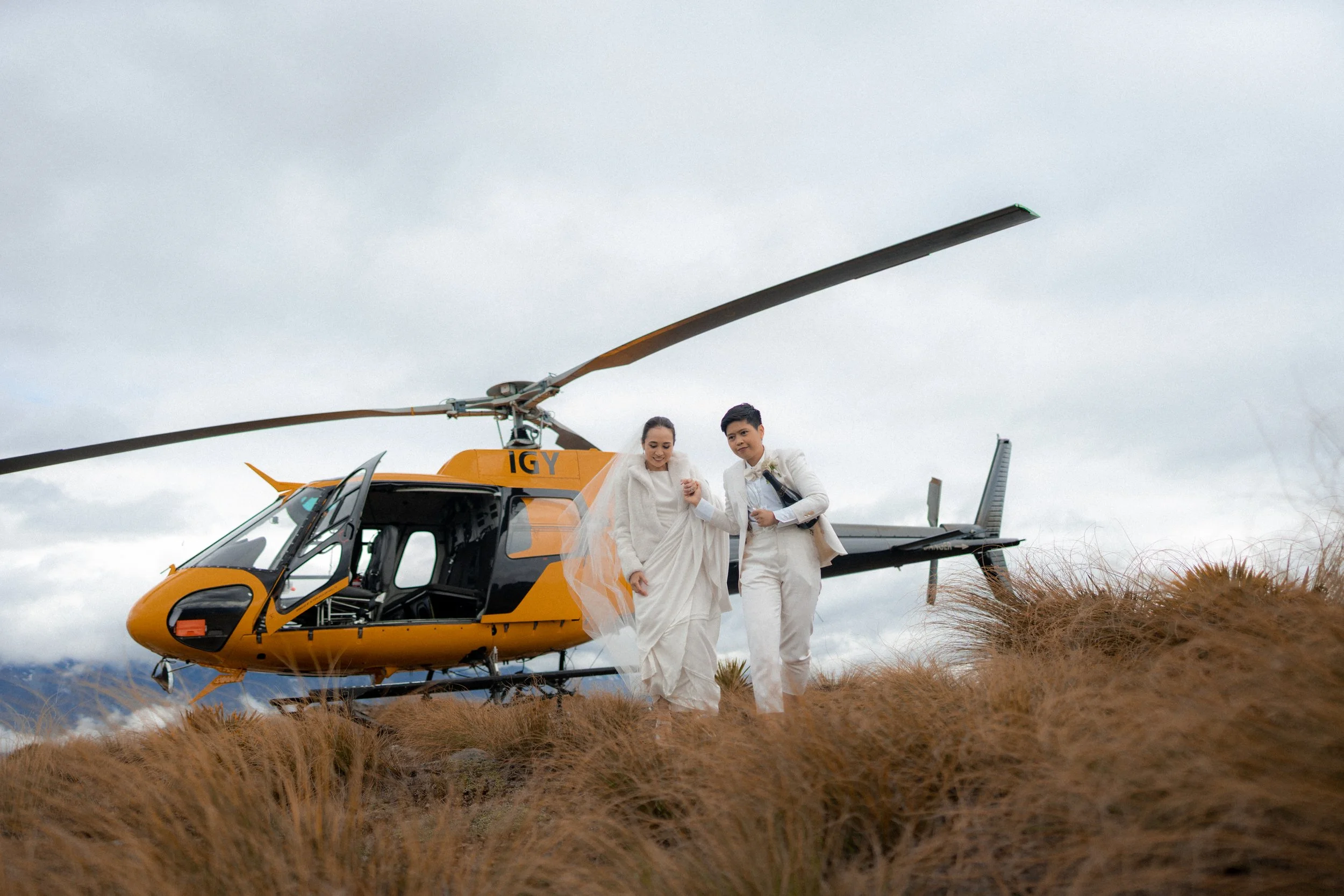 A bride and groom walking hand in hand on grassy terrain with a yellow helicopter in the background under a cloudy sky.