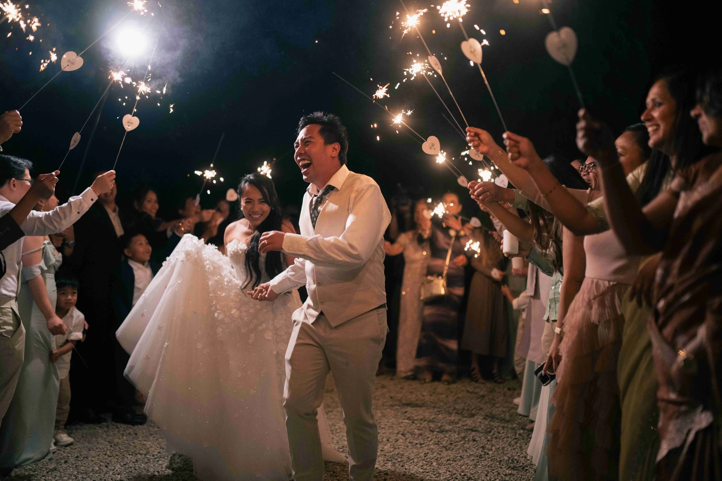 A group of people celebrating outdoors at a wedding at night, holding sparklers. The bride and groom are in the center, smiling and holding hands, with guests surrounding them celebrating.