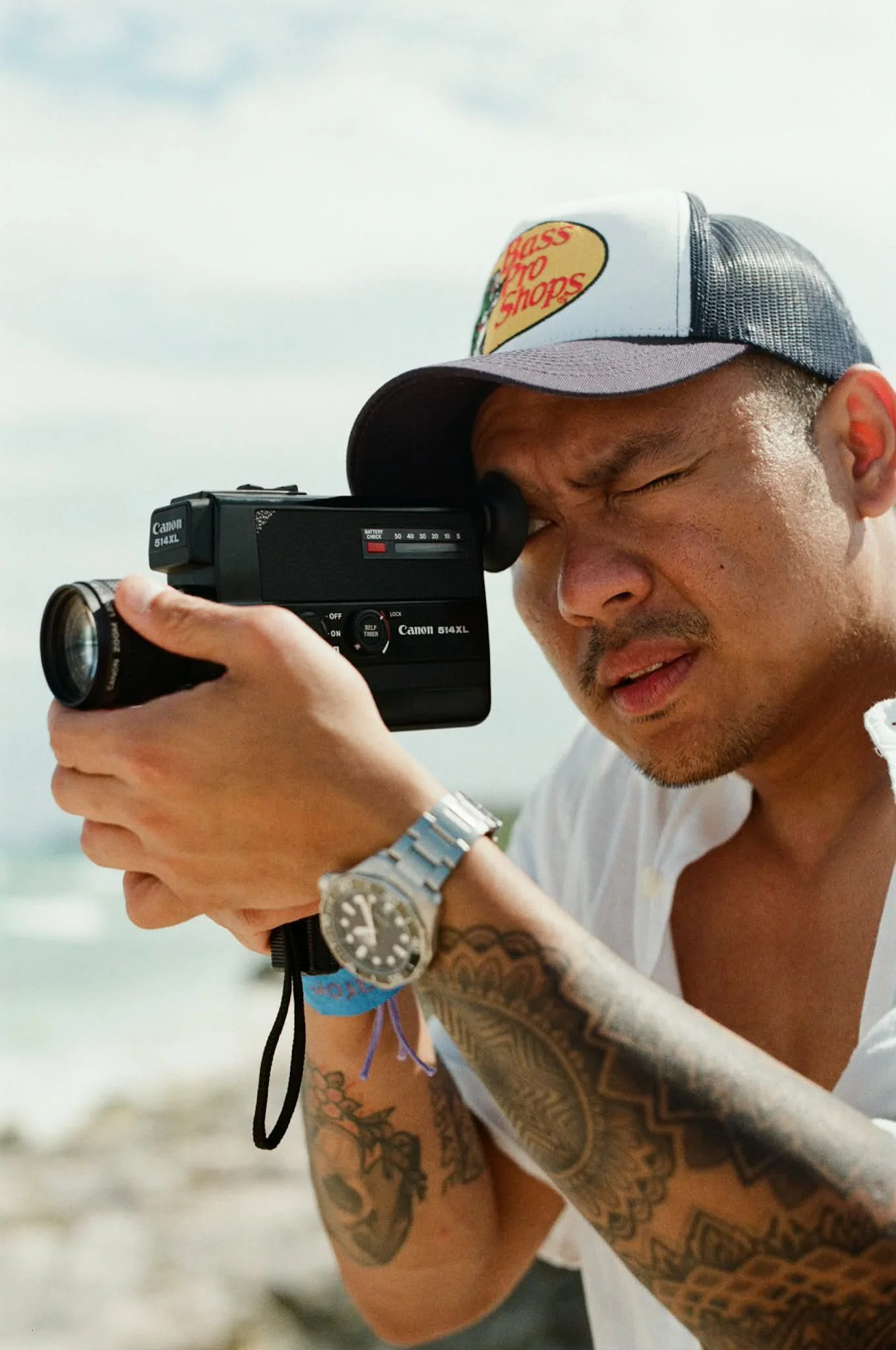 A man with tattoos on his arms looking through the viewfinder of a Canon 514 XL camera. He is wearing a baseball cap with a Patch logo, a white tank top, and a wristwatch.