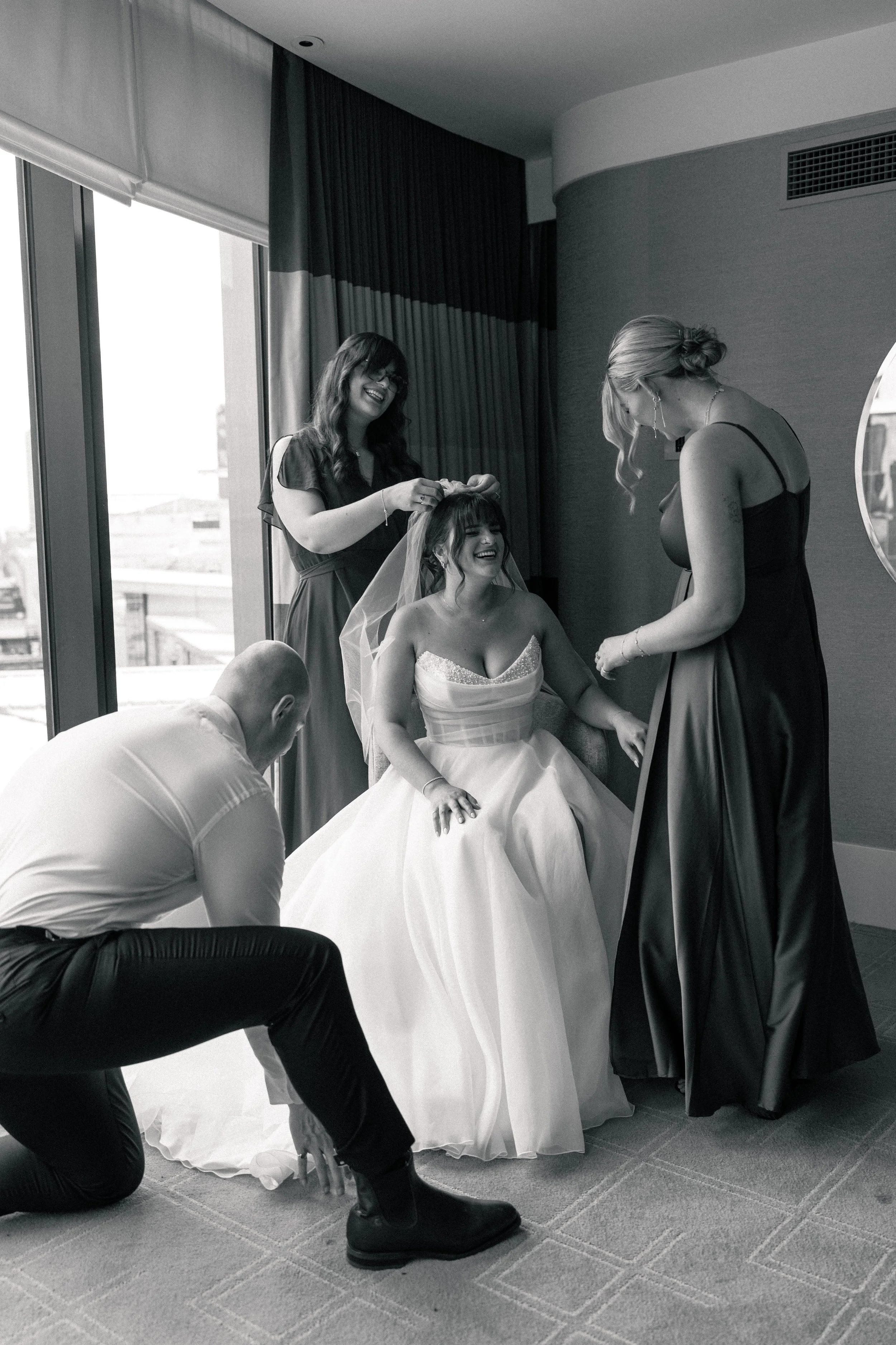 A bride in a wedding dress sitting on a chair, smiling as one woman adjusts her veil and another woman assists her, in a room with large windows and curtains.