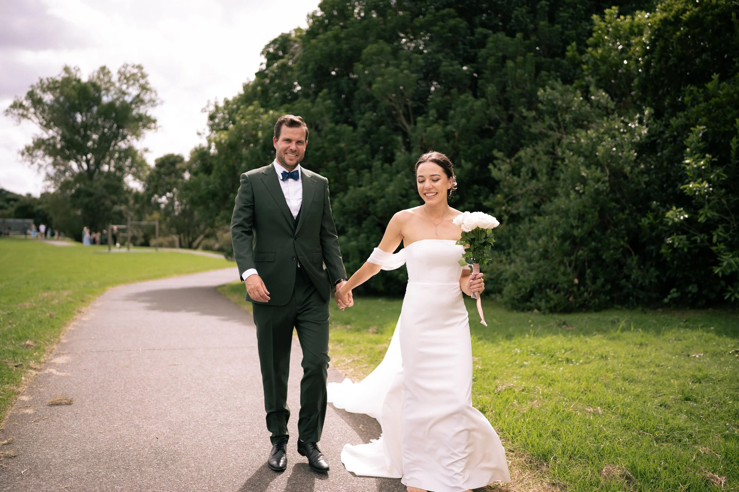 A groom and bride walking hand in hand on a paved path in a park, surrounded by green trees. The groom is wearing a dark suit with a bow tie, and the bride is wearing a white wedding dress holding a bouquet of white roses. They look happy and are smiling.