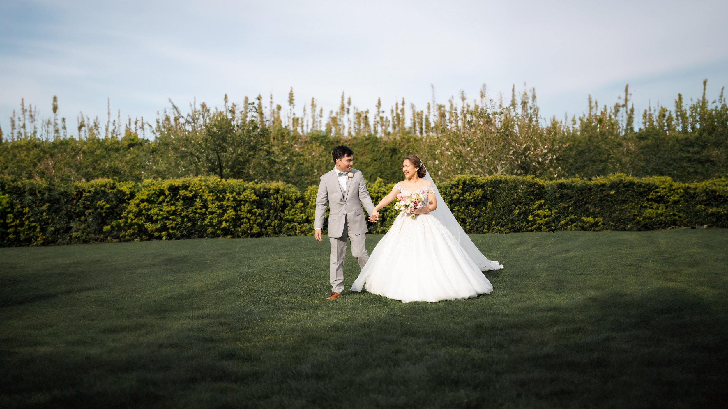 A bride and groom walking hand-in-hand on a grassy lawn with trees and shrubs in the background during a wedding photoshoot.