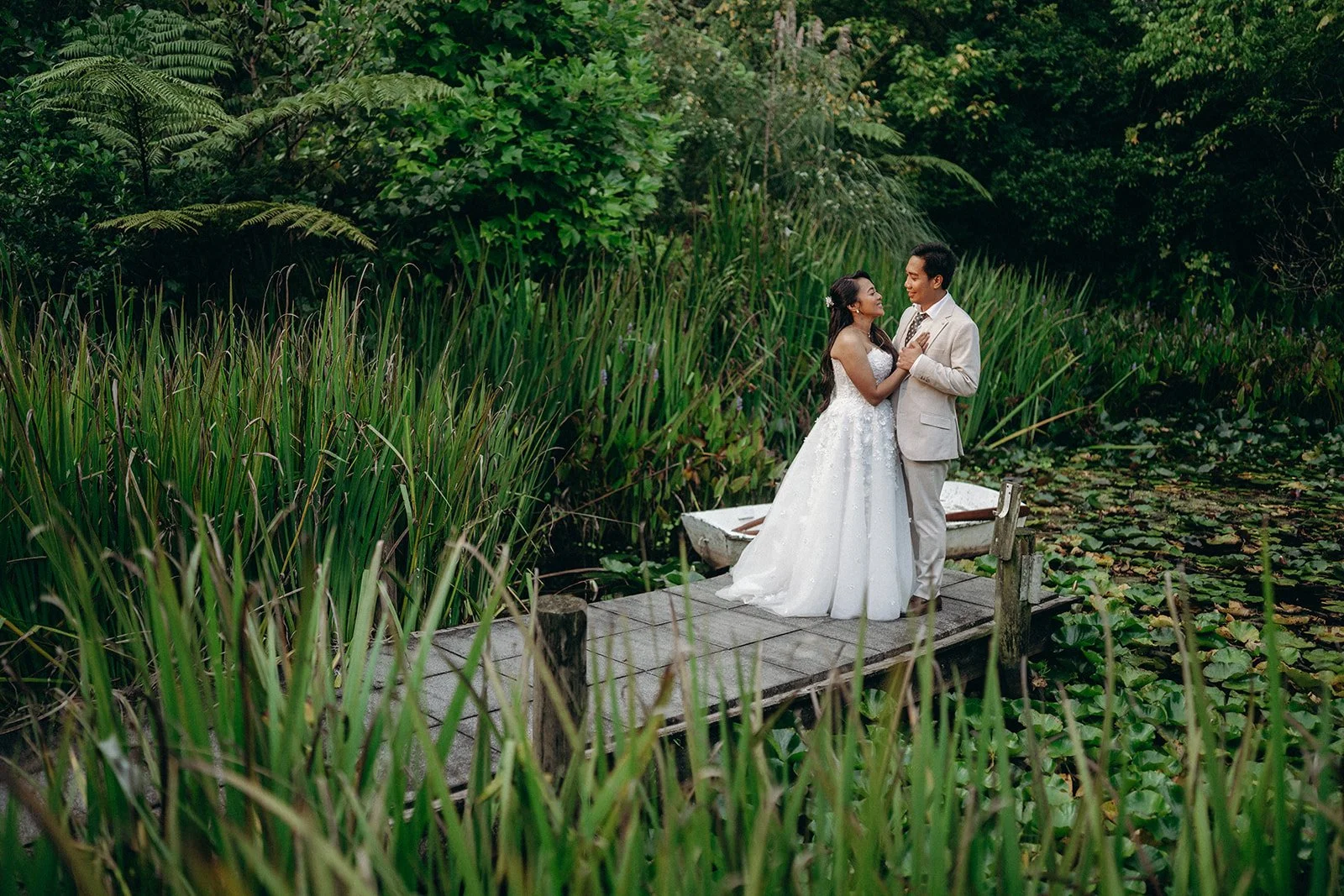 A bride and groom in wedding attire standing on a small wooden dock by a pond, surrounded by lush green plants and trees, sharing a romantic moment.