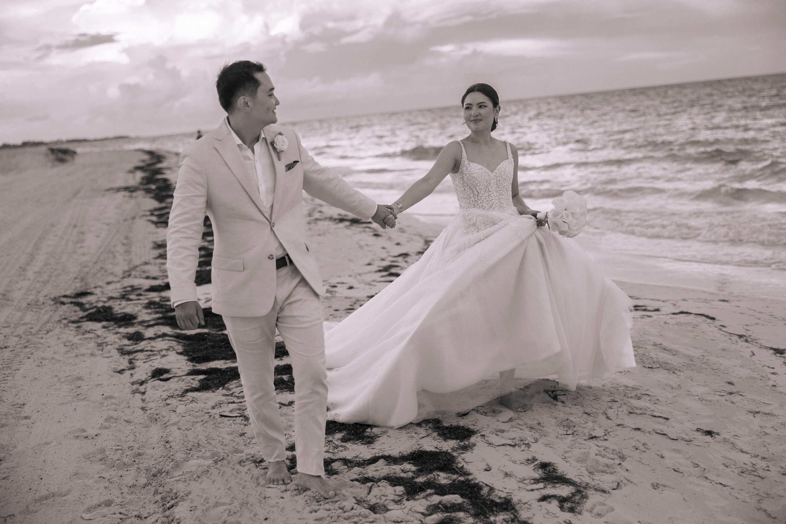 A couple in wedding attire walking hand in hand along a beach at sunset, with the bride holding a bouquet of flowers.