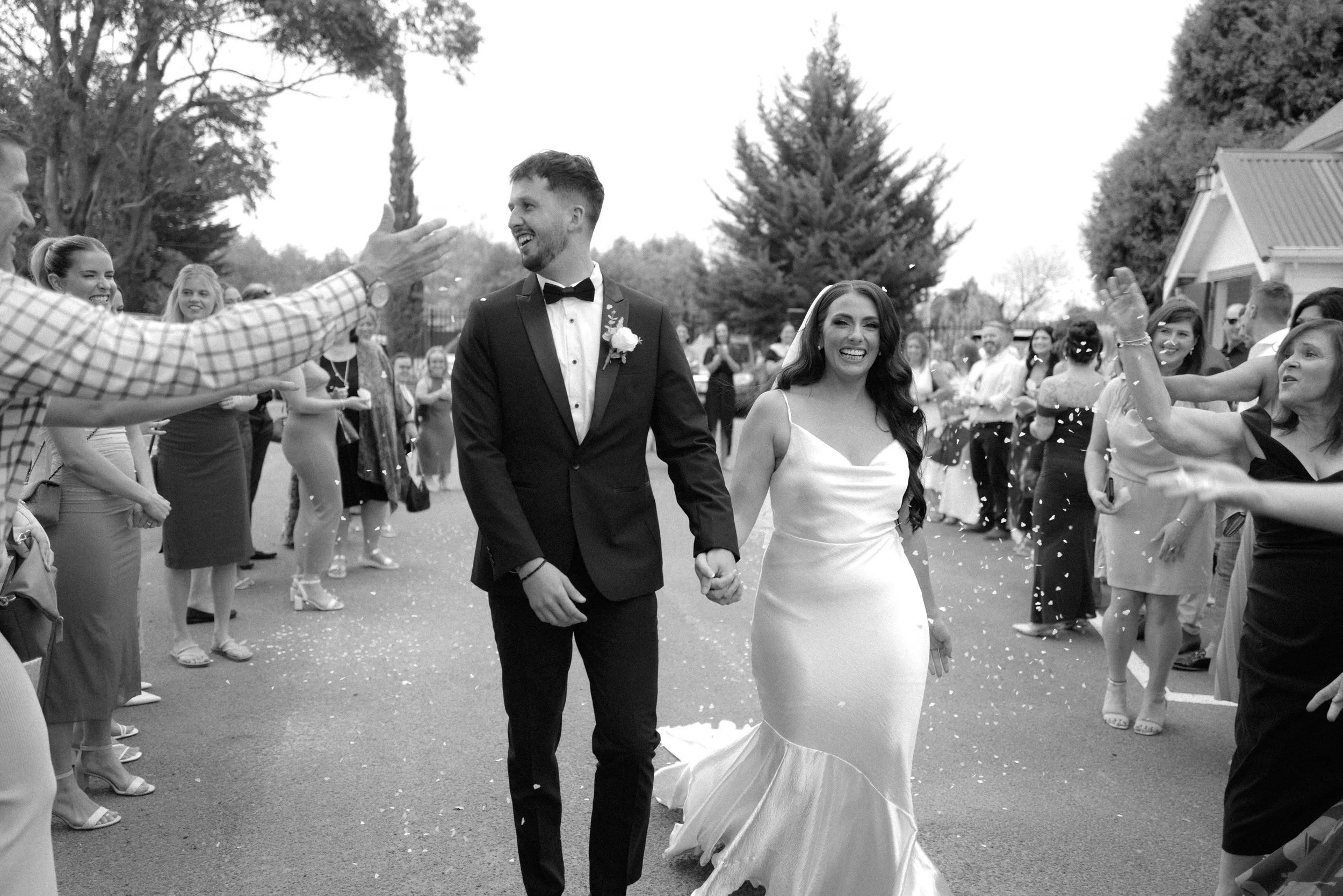 Black and white photo of a newlywed couple walking hand in hand during their outdoor wedding reception, surrounded by friends and family celebrating and throwing confetti.