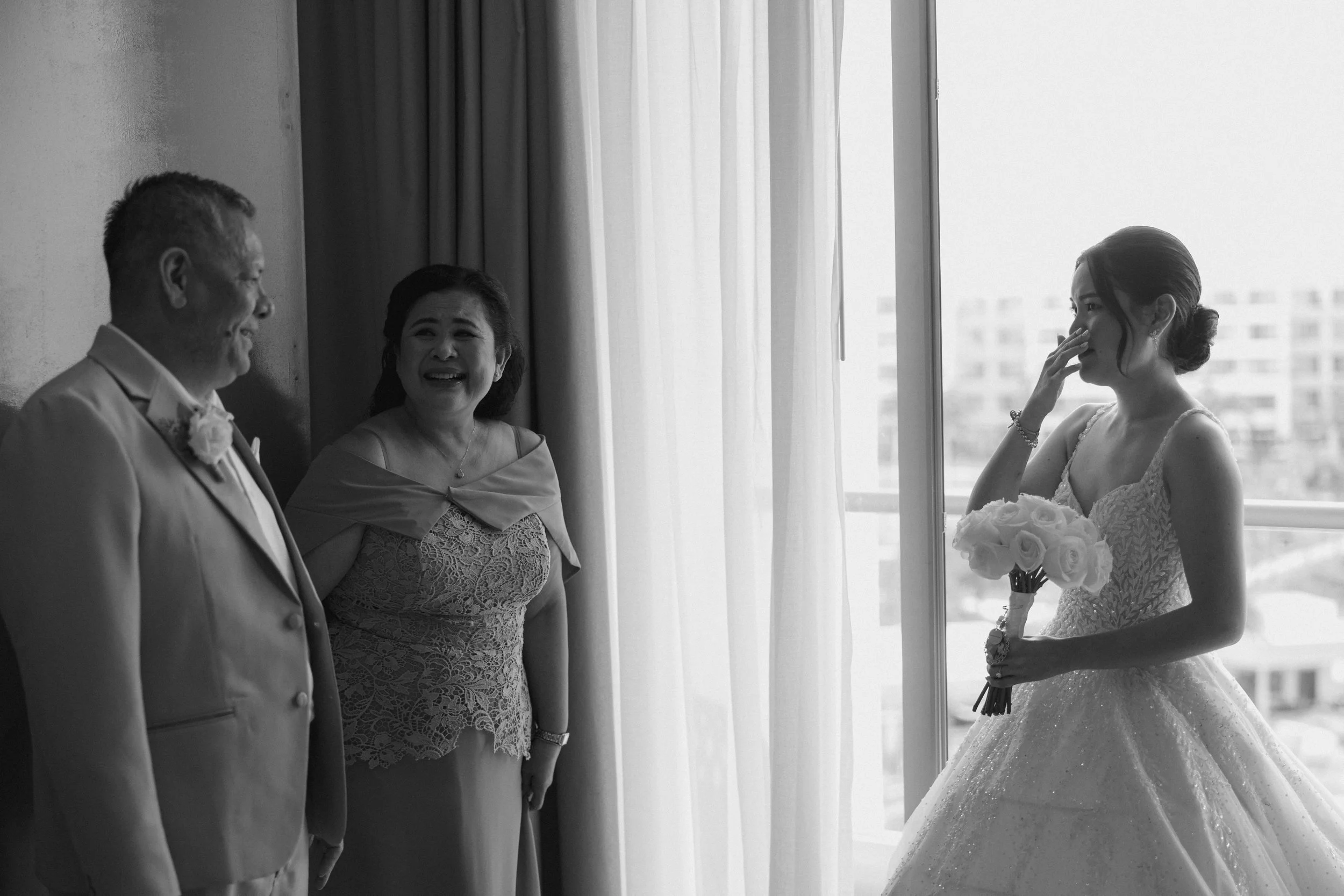 A black and white photo of a wedding scene showing a bride holding a bouquet of flowers looking at her father and mother, who are smiling at her. They are standing indoors near a large window.