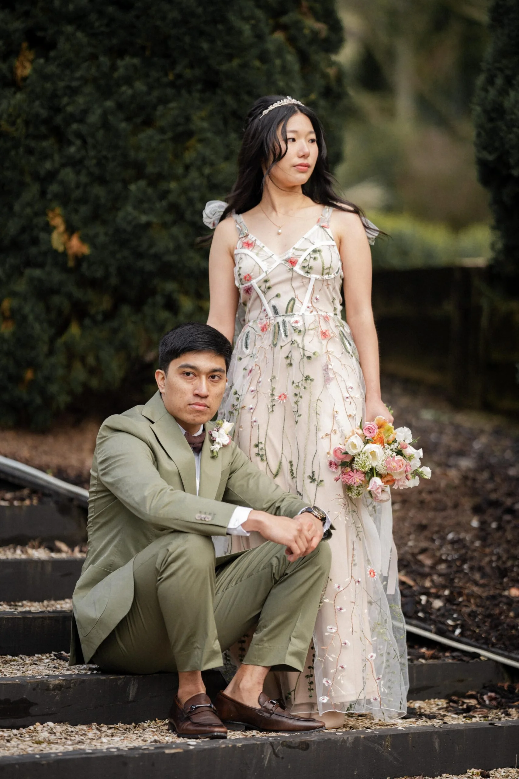 A young couple dressed in formal wedding attire outdoors, with the woman holding a bouquet of flowers while the man sits on steps, surrounded by lush green foliage.