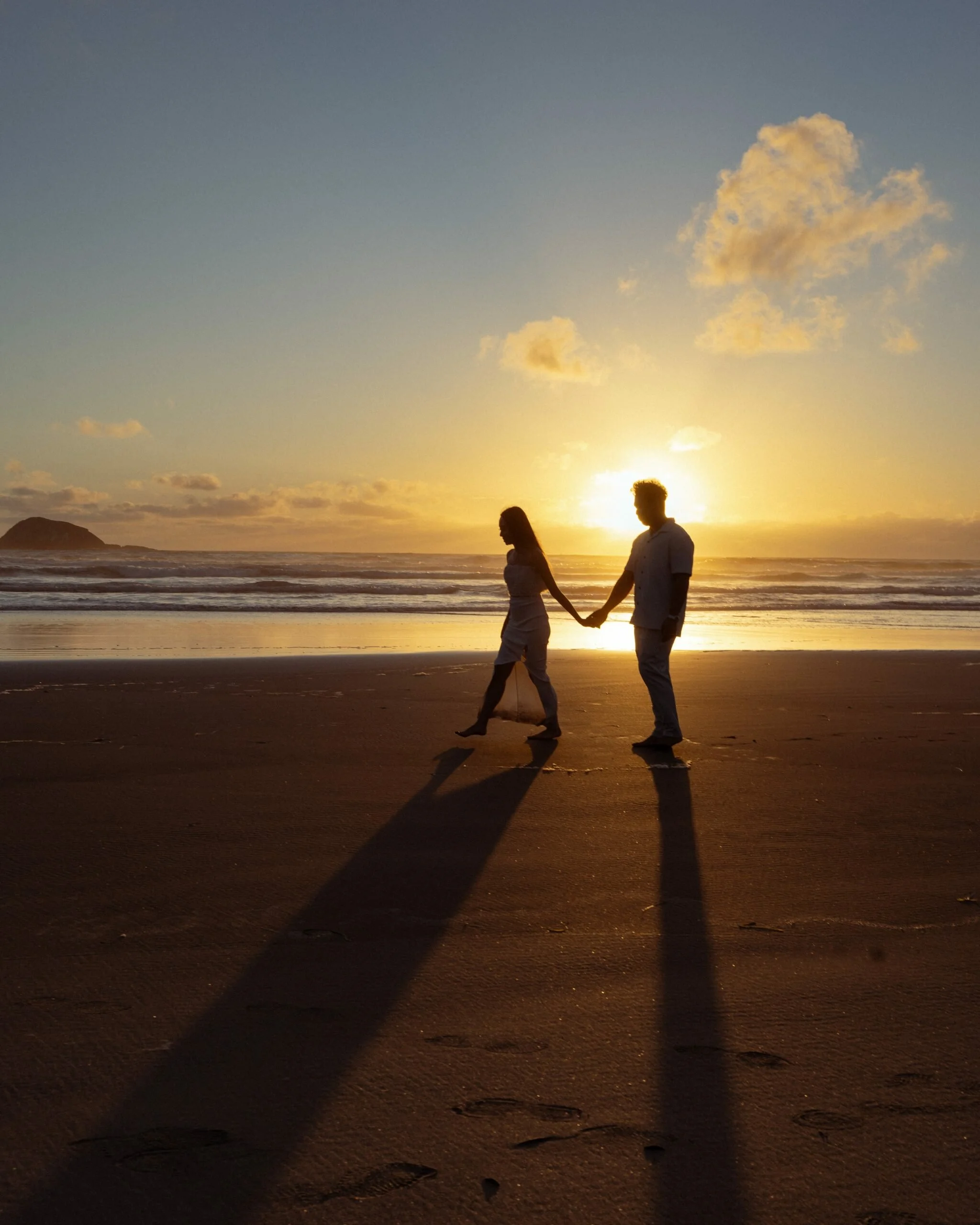 A silhouetted couple holding hands on a beach at sunset with long shadows and cloud-filled sky.