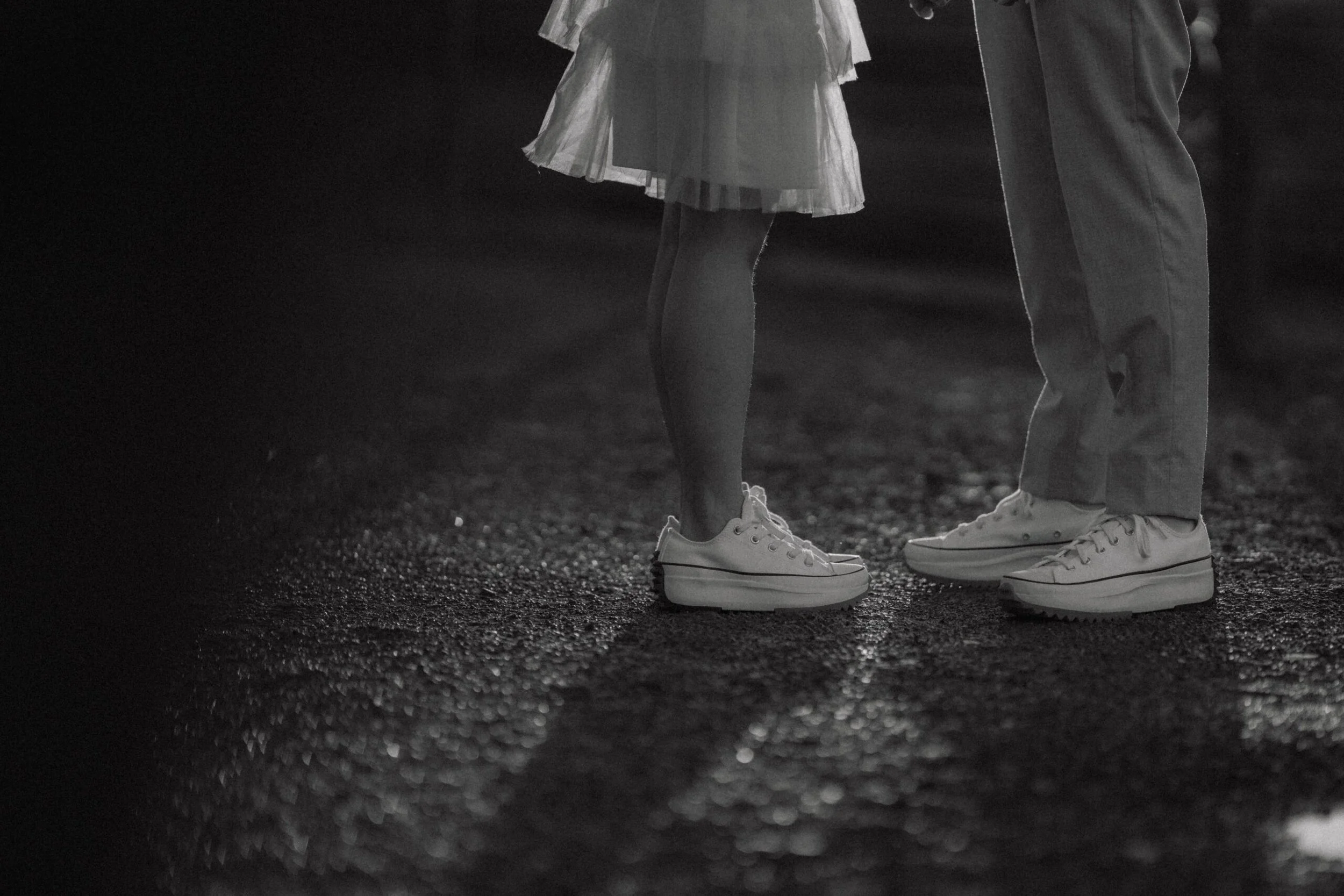 A young girl and an adult man holding hands on a dark street at night, both wearing white sneakers, with the girl in a dress and the man in pants.