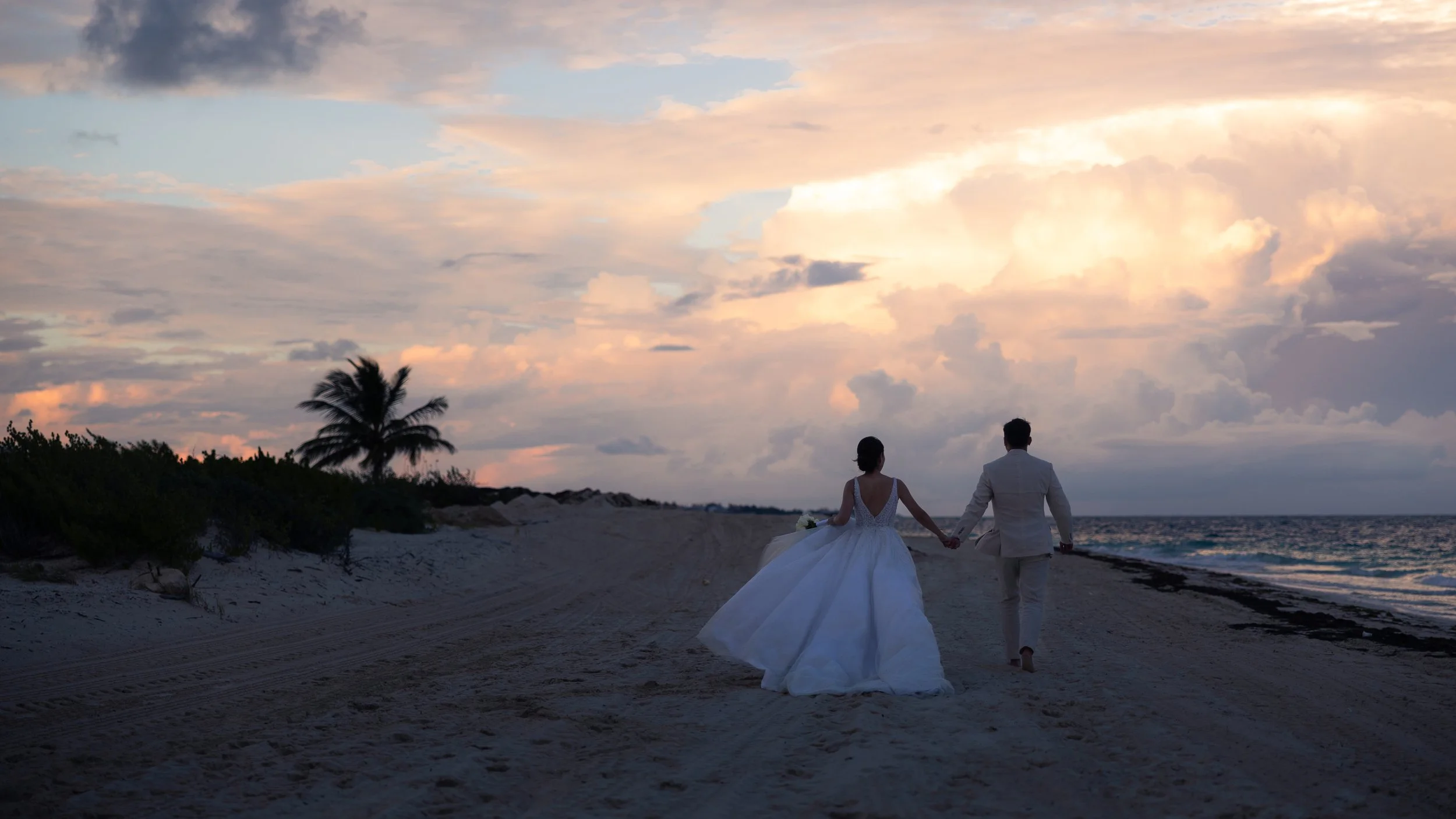 A bride and groom walking hand in hand along the beach during sunset, with clouds in the sky and a palm tree in the background.