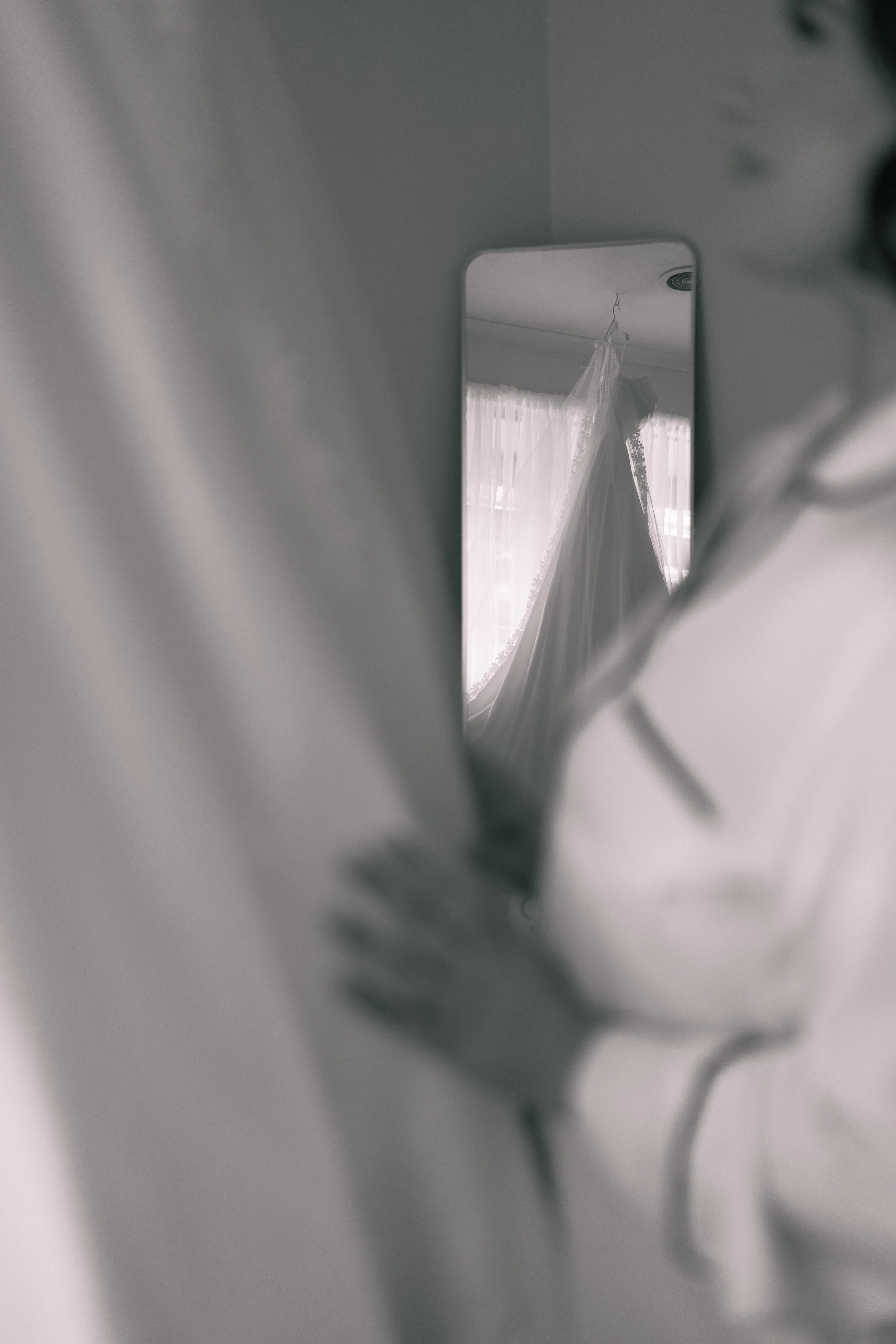 A blurred foreground with a mirror reflecting a wedding dress hanging in front of a window with curtains.