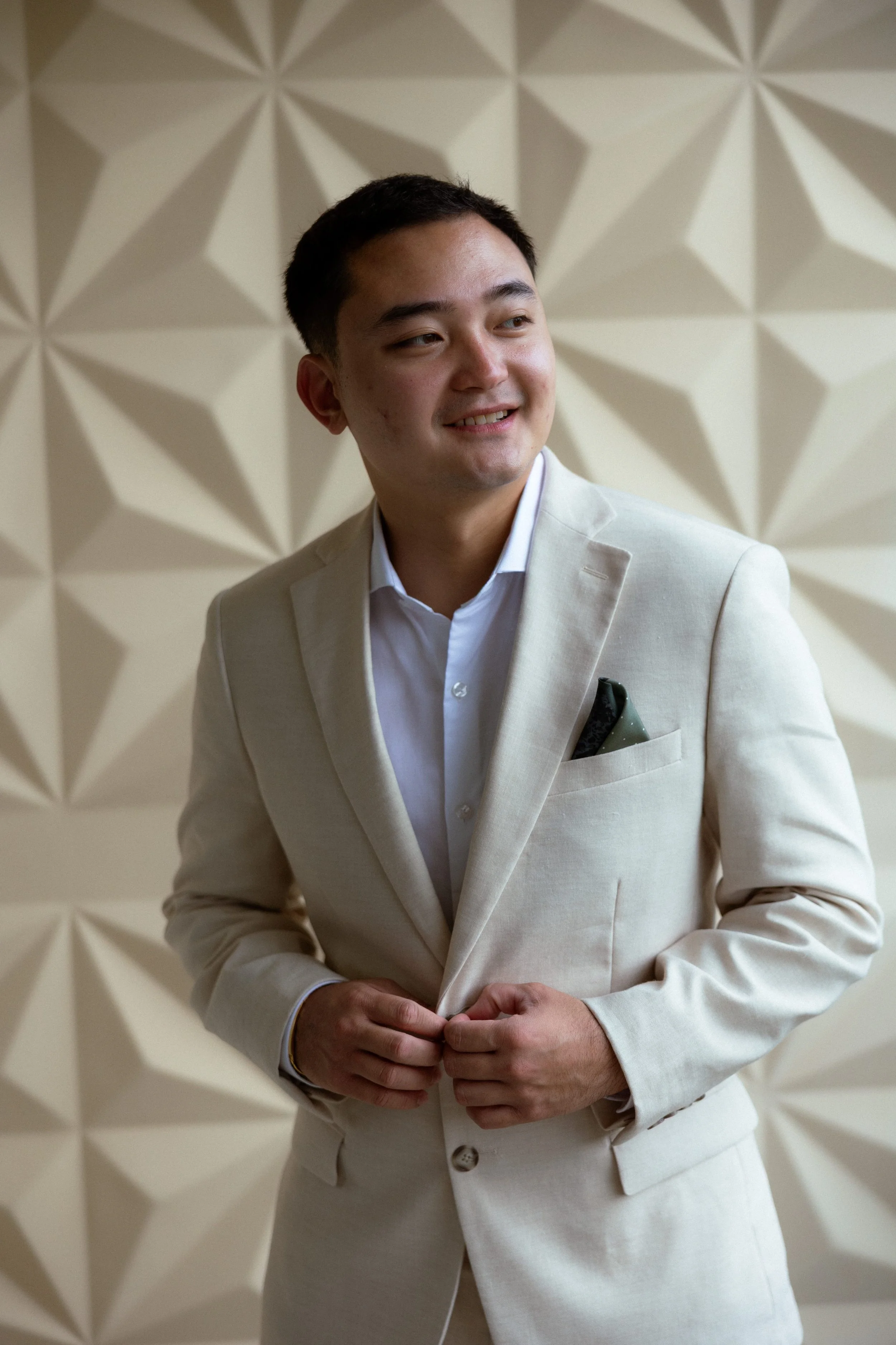 A man in a light-colored suit with a white shirt standing against a 3D geometric patterned wall.