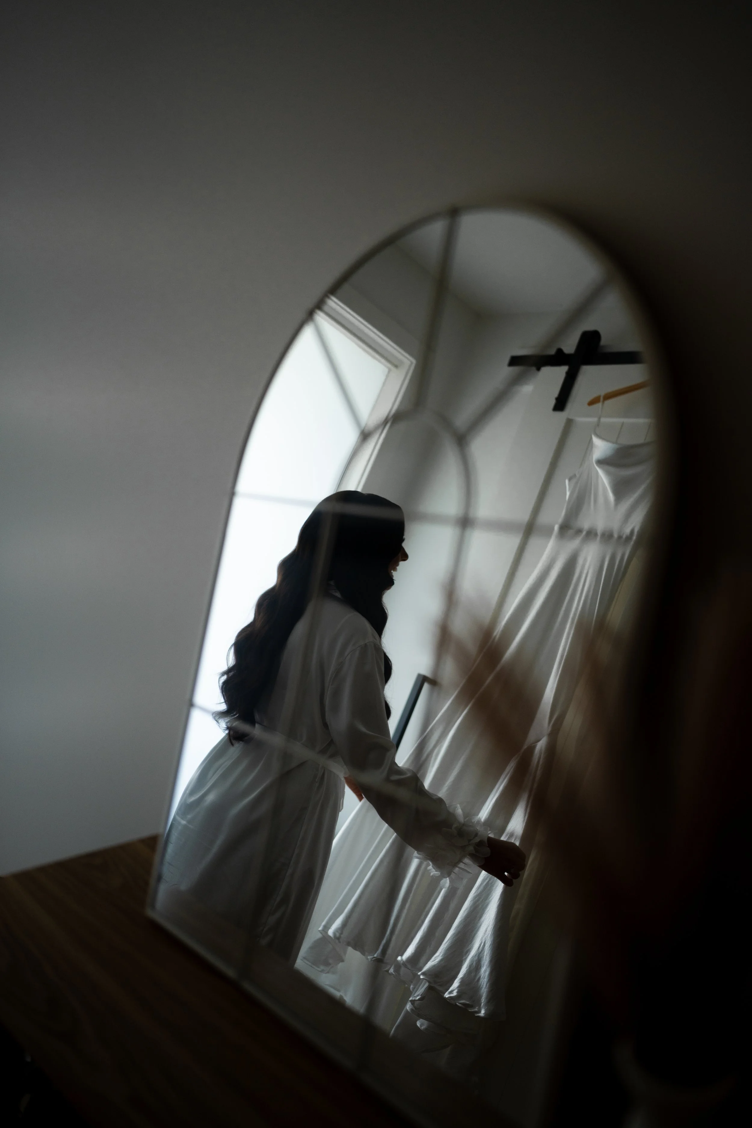Reflection of a woman in a mirror as she looks at a white dress hanging on a rack in a well-lit room.