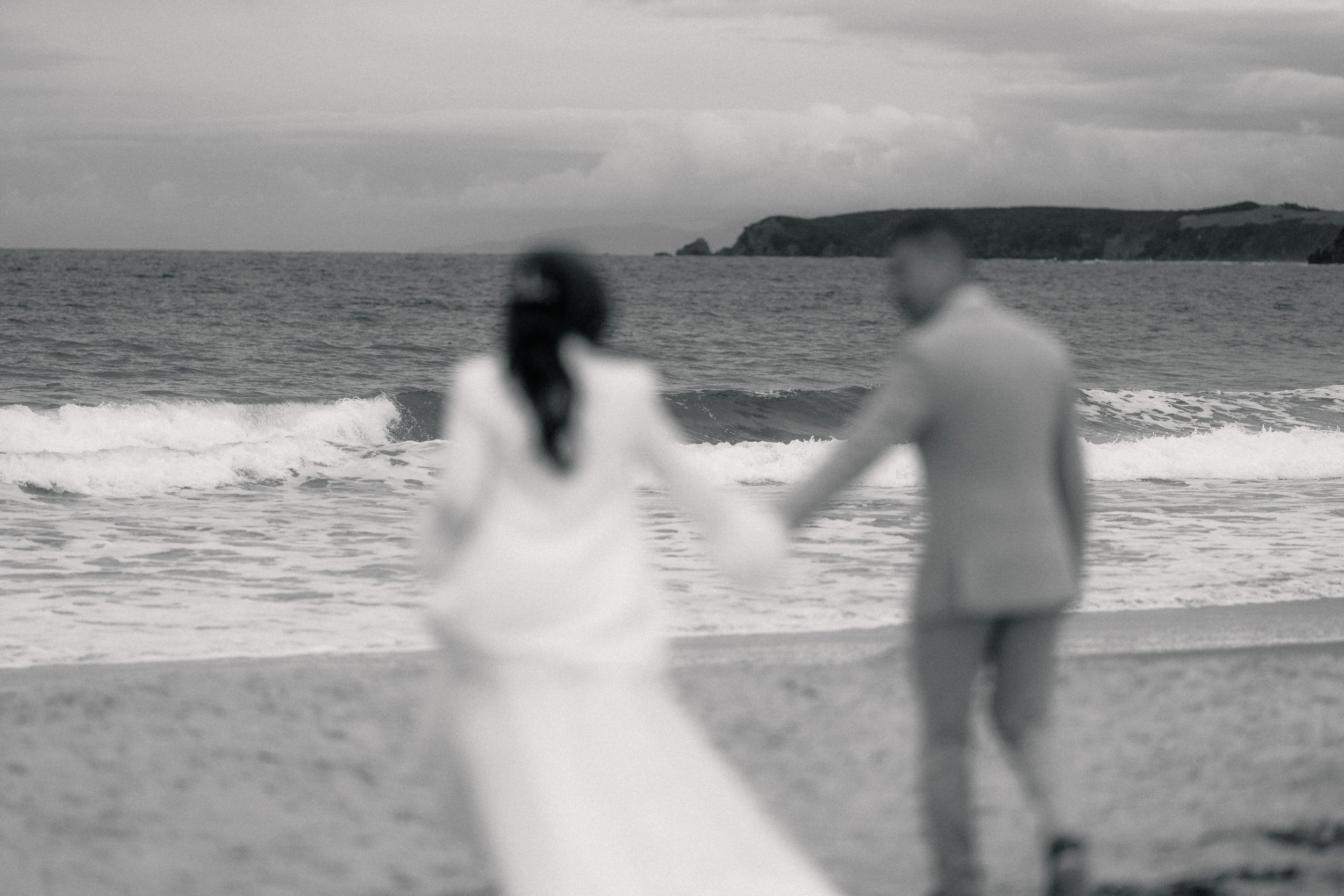 A couple holding hands on a beach, with the ocean and clouds in the background, in black and white.