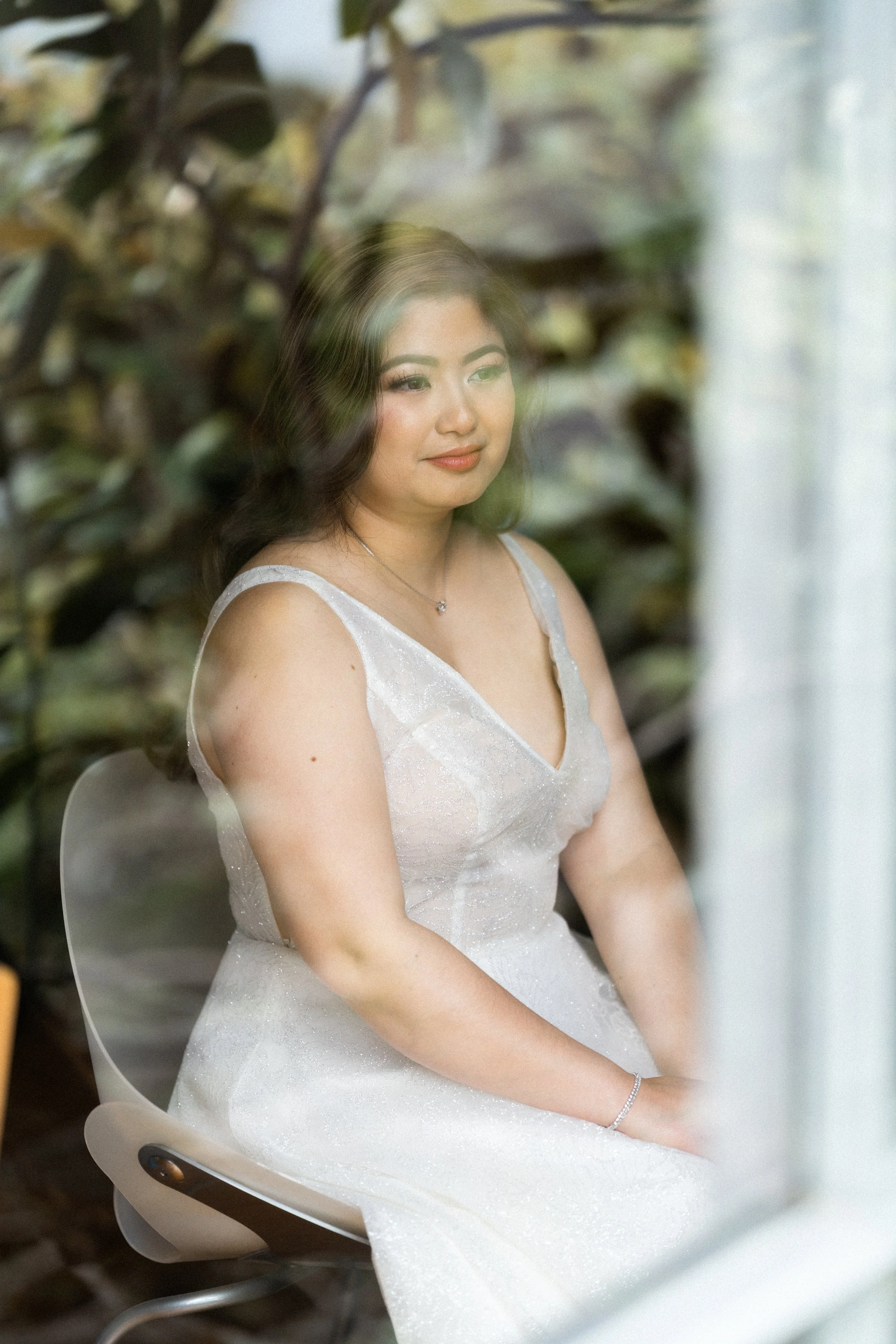 A woman in a white dress sitting on a modern chair, looking thoughtfully through a window with a blurred garden background.