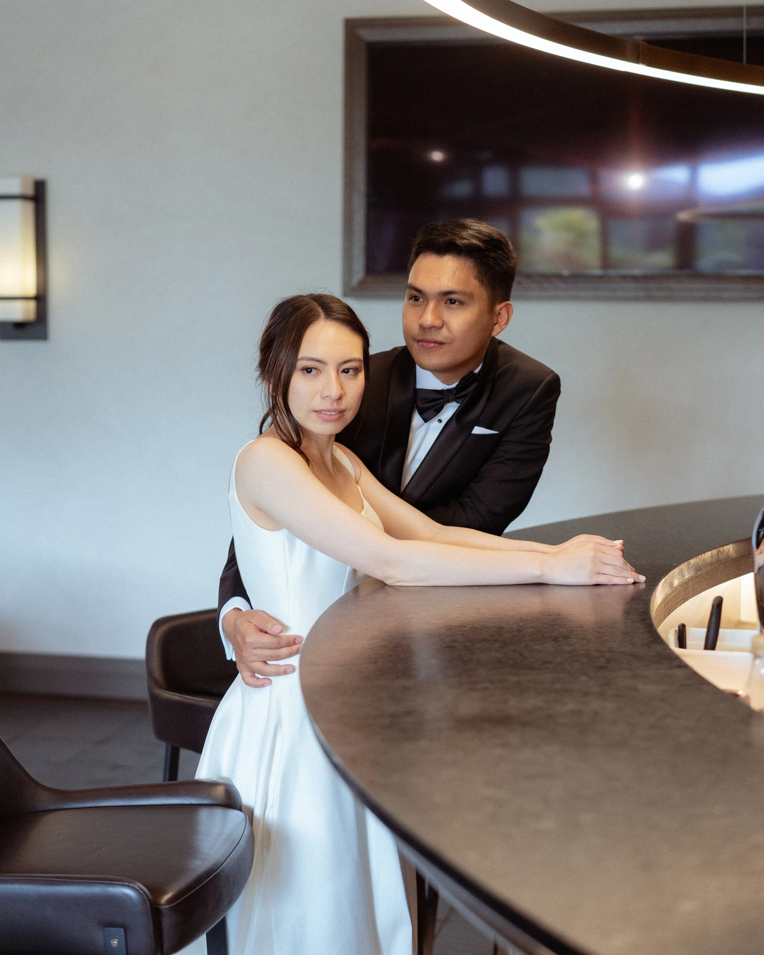 A newlywed couple in formal attire, the woman in a white wedding dress and the man in a black tuxedo, are posing at a bar counter in an indoor setting.