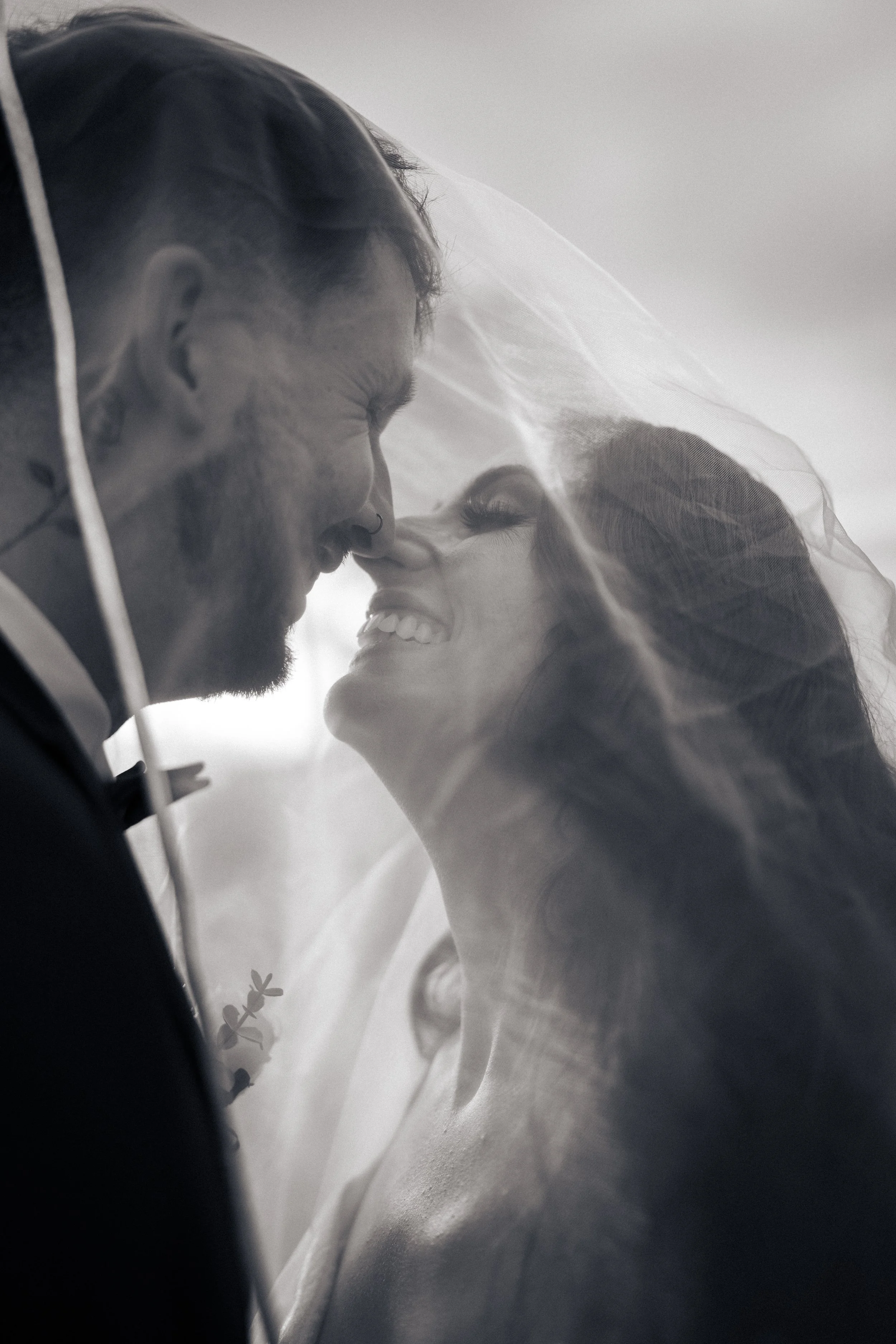 A black-and-white photo of a bride and groom with their noses touching, smiling under a veil.