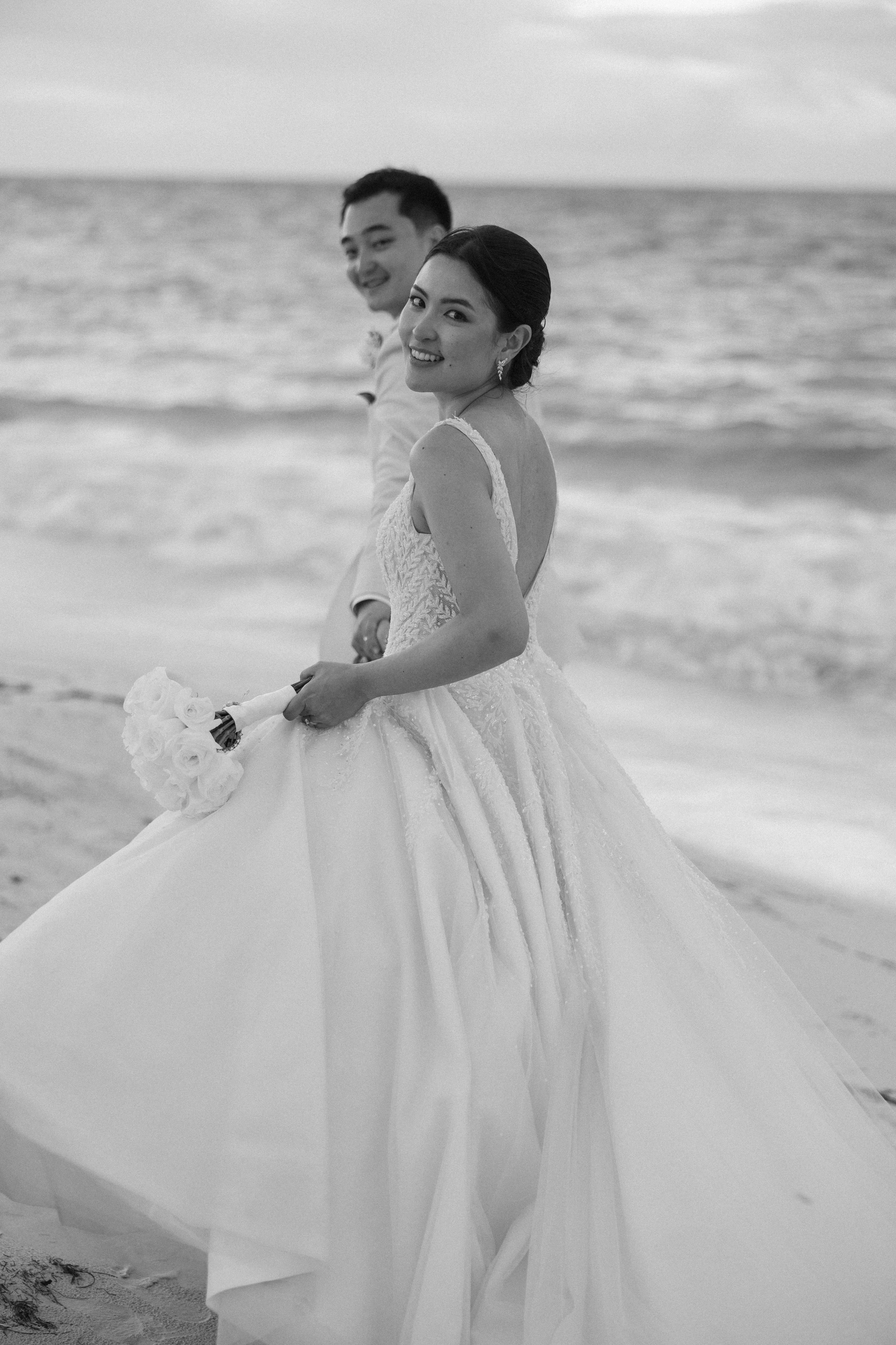 Black and white photo of a bride in a wedding gown holding a bouquet of flowers, standing on a beach with the ocean in the background. Behind her is a groom in formal attire.