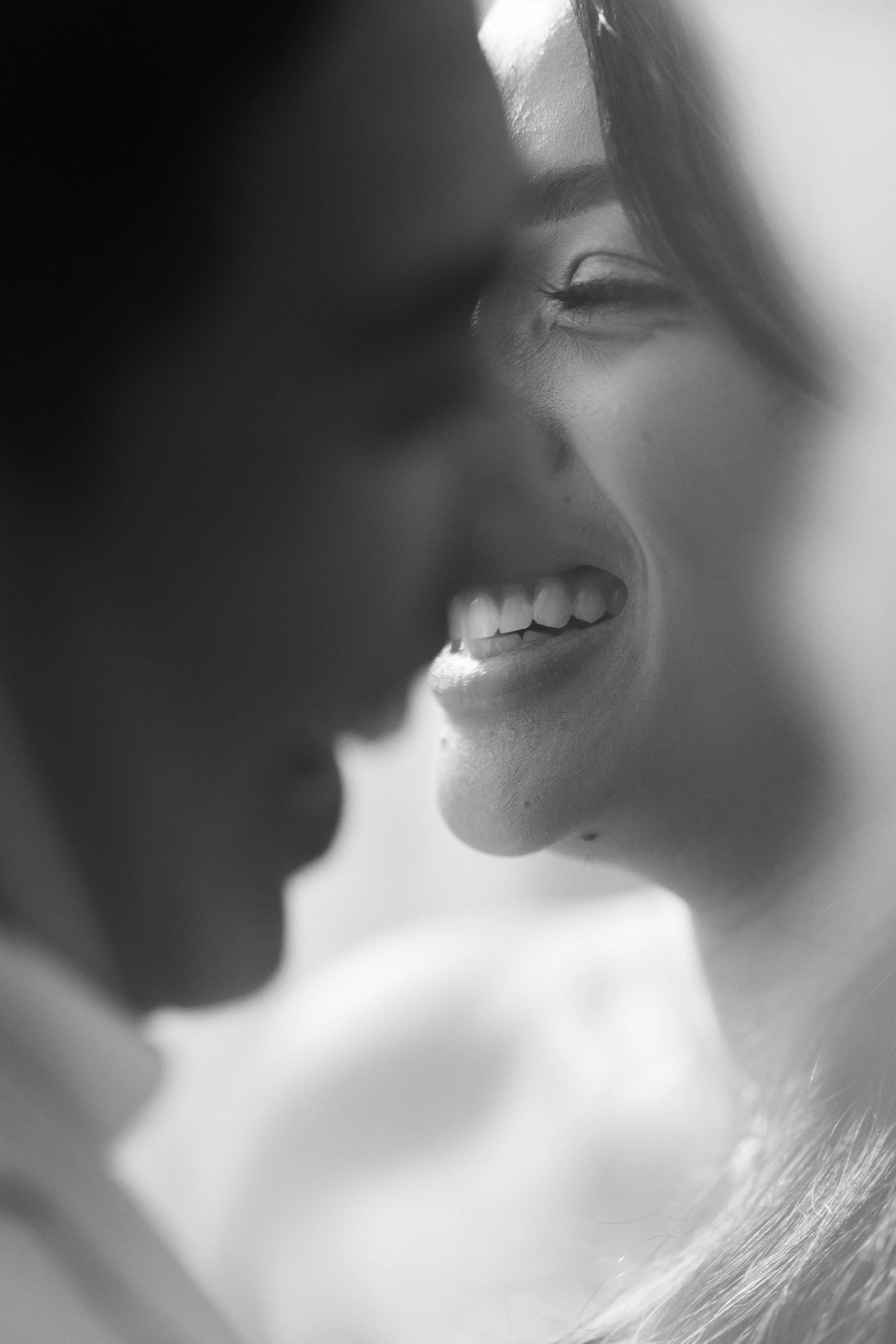 Close-up black and white photo of a woman smiling with eyes closed, facing another person, in a tender moment.