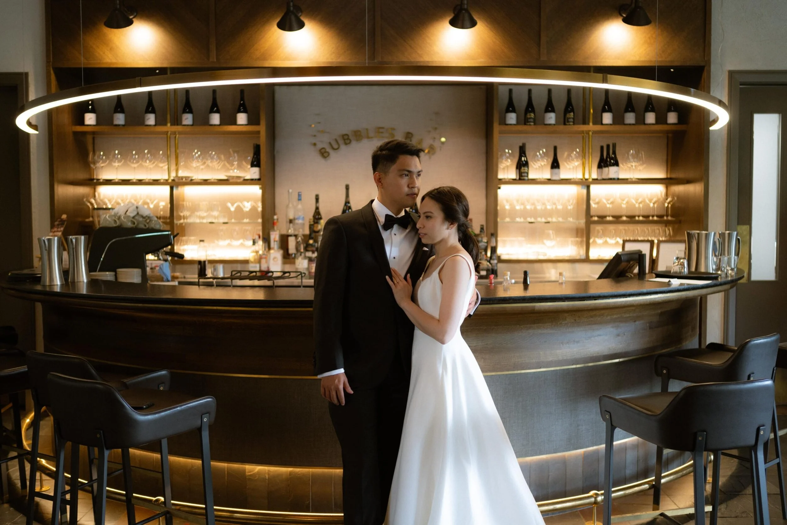 A newlywed couple, a man in a black tuxedo and woman in a white wedding dress, standing close and embracing at a bar or lounge area.