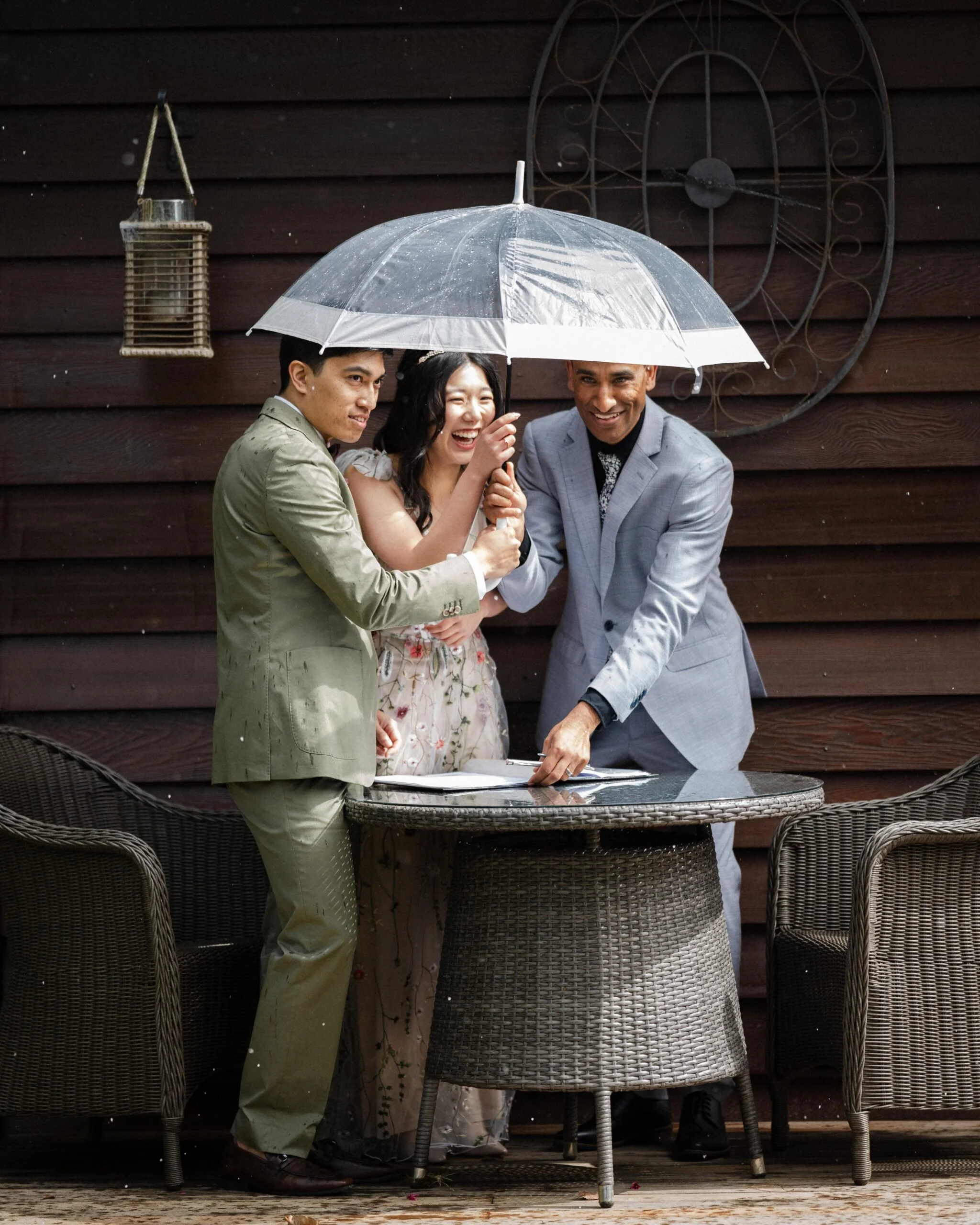 Three people standing under an umbrella around a table, smiling and holding hands, in front of a wooden wall.