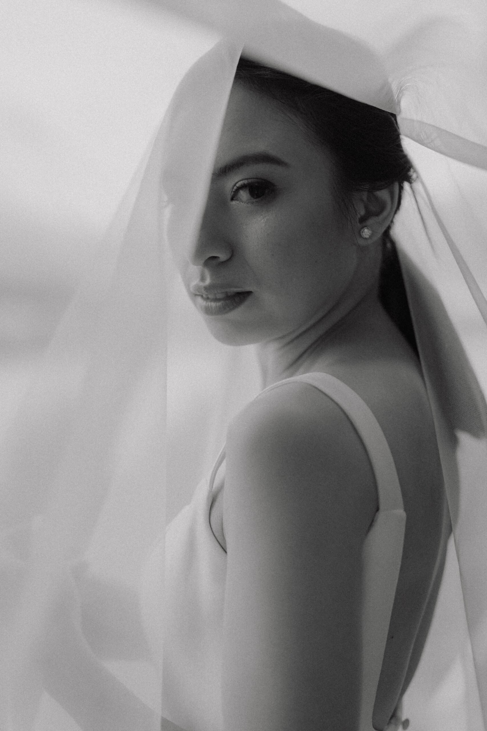 A black and white portrait of a woman with a veil over her face, looking over her shoulder at the camera.