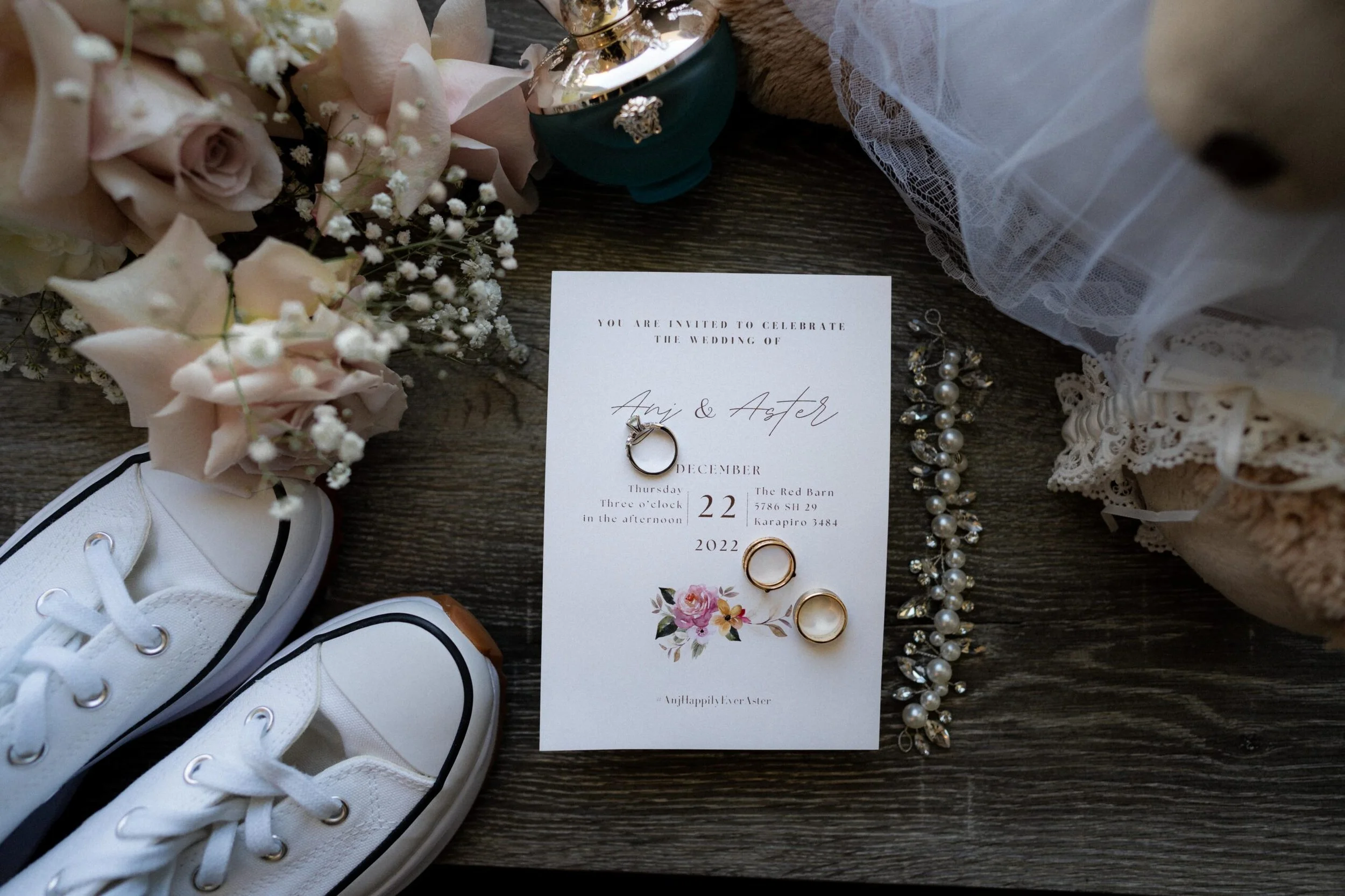 Wedding invitation, rings, jewelry, sneakers, flowers, and lace fabric on a wooden surface.