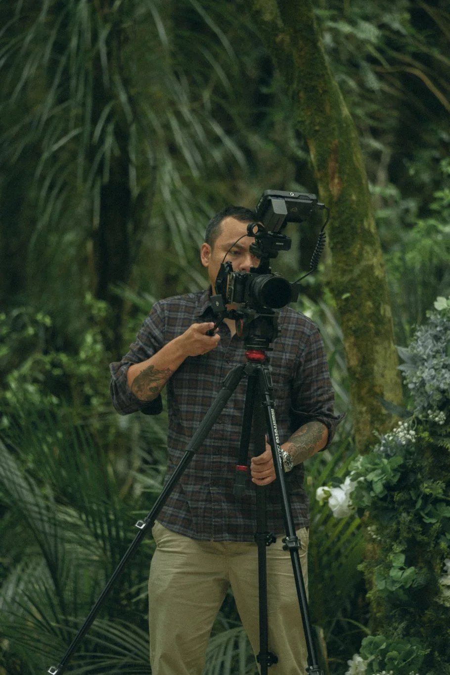 A man operating a professional camera on a tripod in a lush, green outdoor setting with plants and trees.