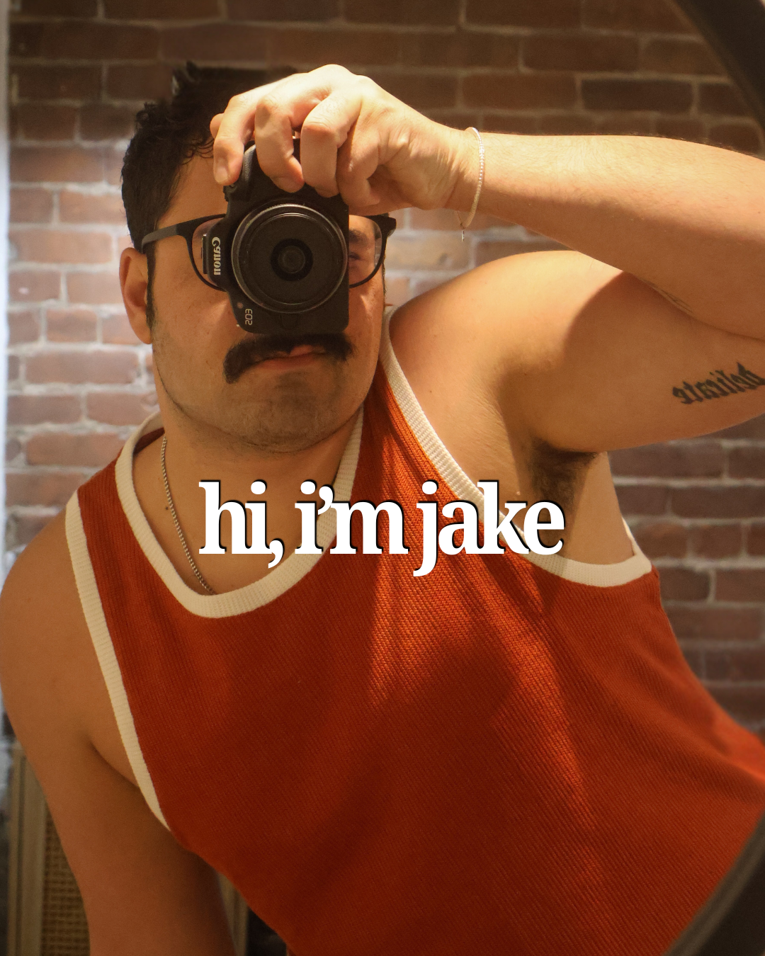 A man taking a selfie in front of a mirror with a brick wall background, wearing glasses, a mustache, a red sleeveless top, and a silver chain around his neck. The caption says, 'hi, im jake.'