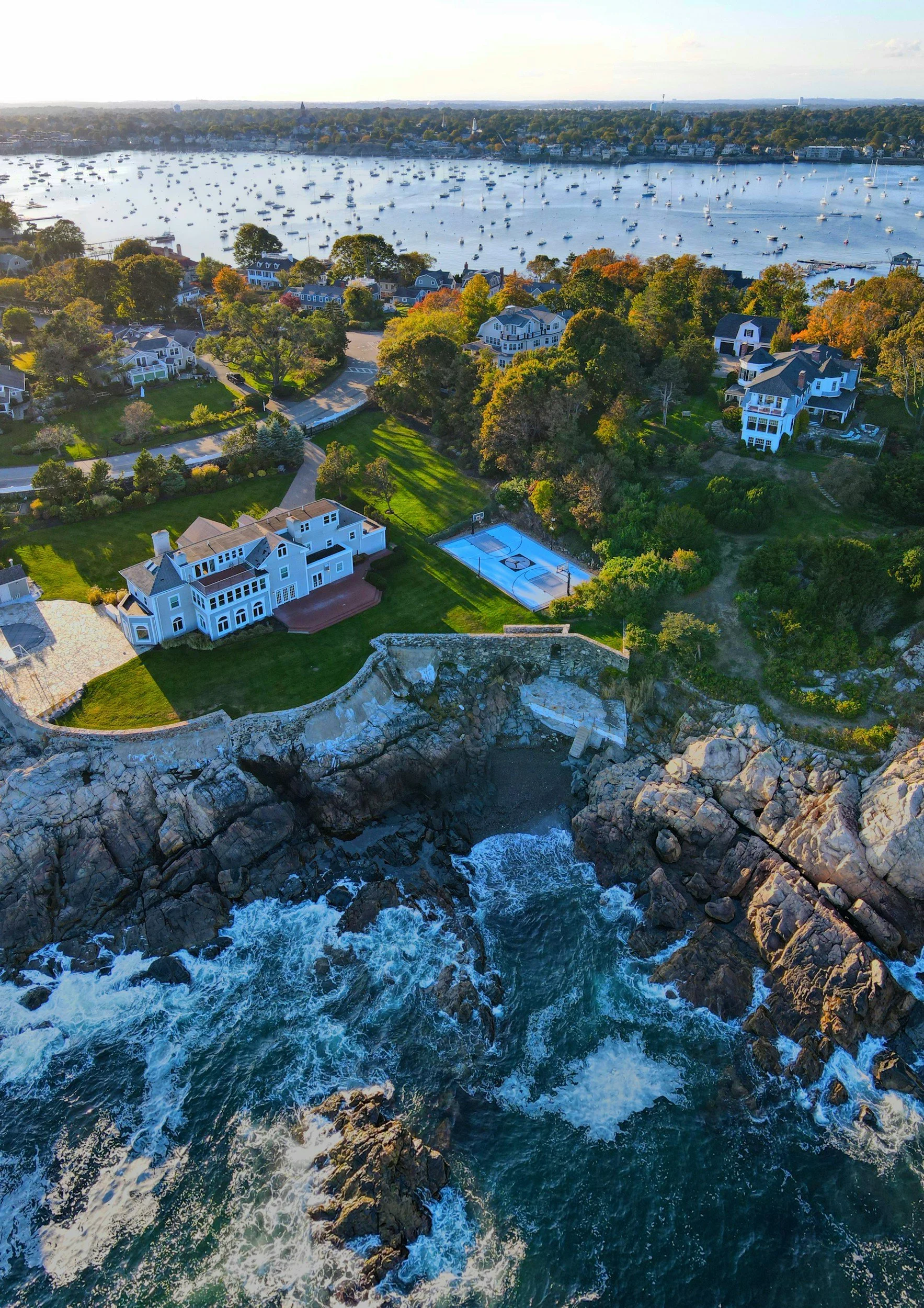Aerial view of a coastal neighborhood with houses, trees, a basketball court, and a large body of water filled with sailboats.