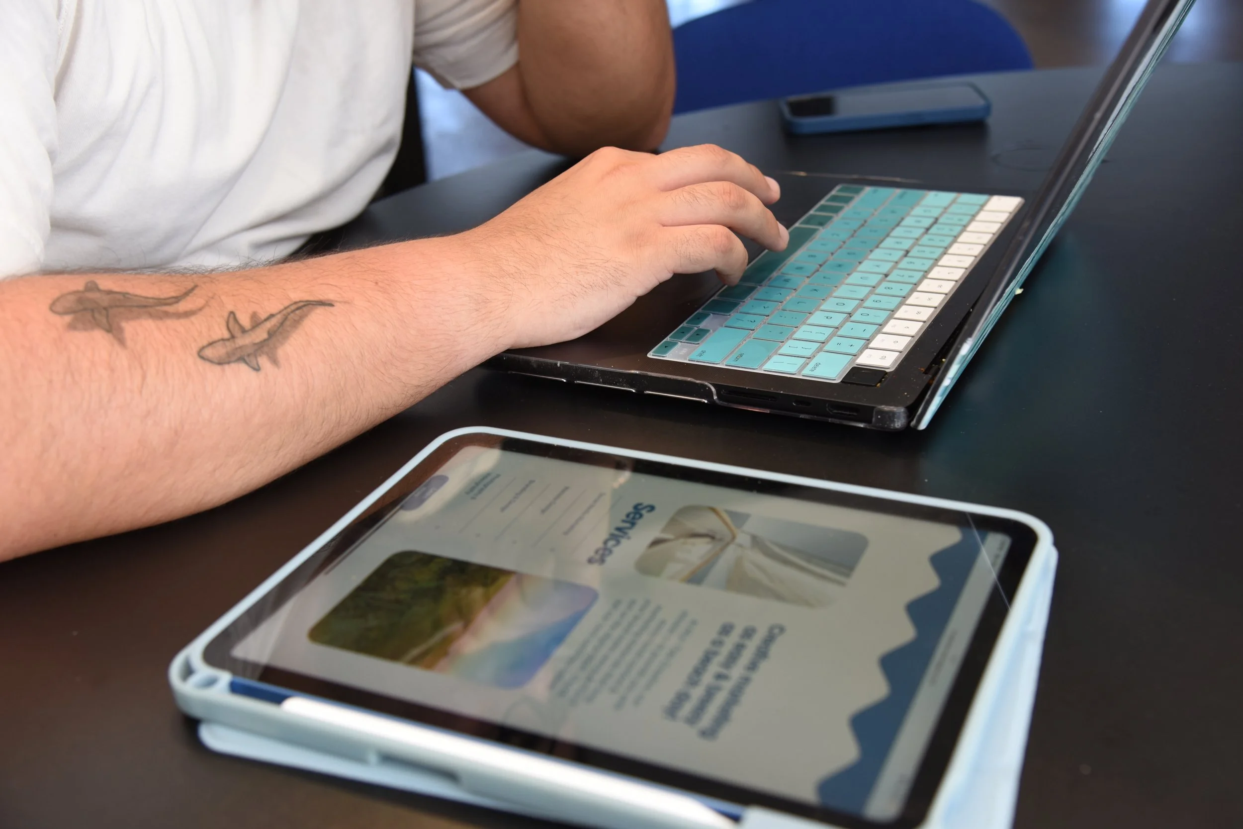 A person sitting at a table using a laptop, with a smartphone nearby, and a tablet displaying a webpage in the foreground.