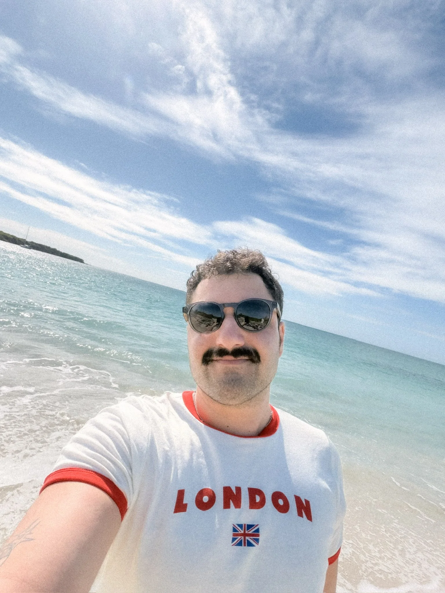 A man wearing sunglasses and a white t-shirt with red trim and the words 'LONDON' and a Union Jack flag tattooed on his arm is taking a selfie on the beach with blue water and a cloudy sky in the background.