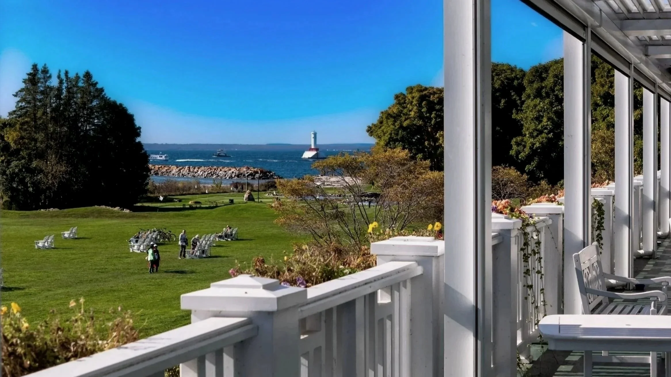 View from the balcony at Mission Point Resort overlooking a lawn with trees, people, a lighthouse, boats in the water, and a clear blue sky.