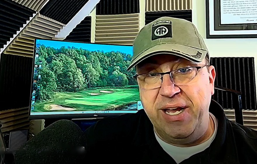 The regional podcast host wearing glasses, a baseball cap, and a black jacket speaking into a microphone in front of a computer monitor displaying a golf course with trees and sand traps.