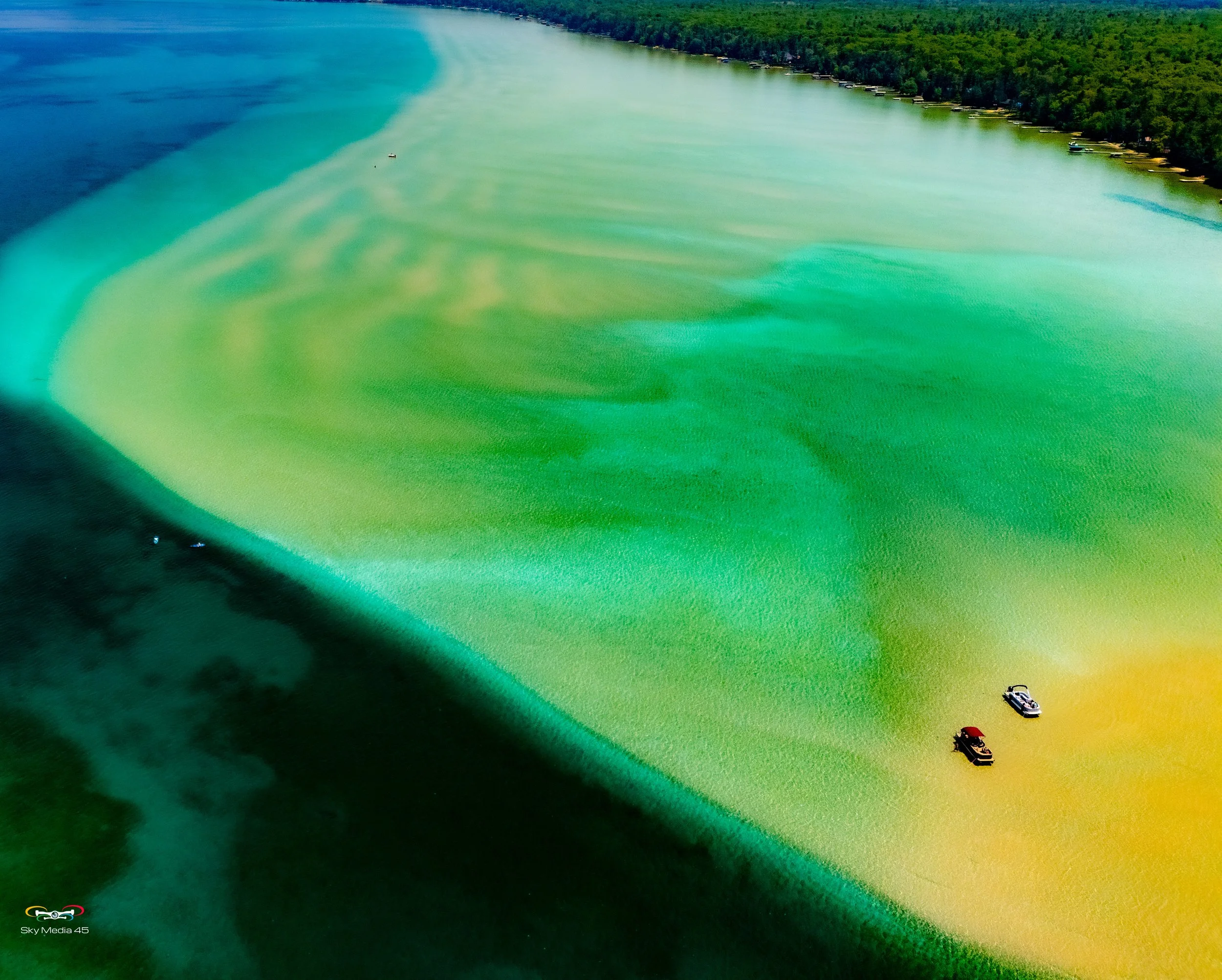 An aerial view of the iconic Torch Lake sandbar with a gradient of colors from dark blue to bright green, bordered by green forested land on one side. Two cars are parked on the yellow sandy shore at the bottom right corner.