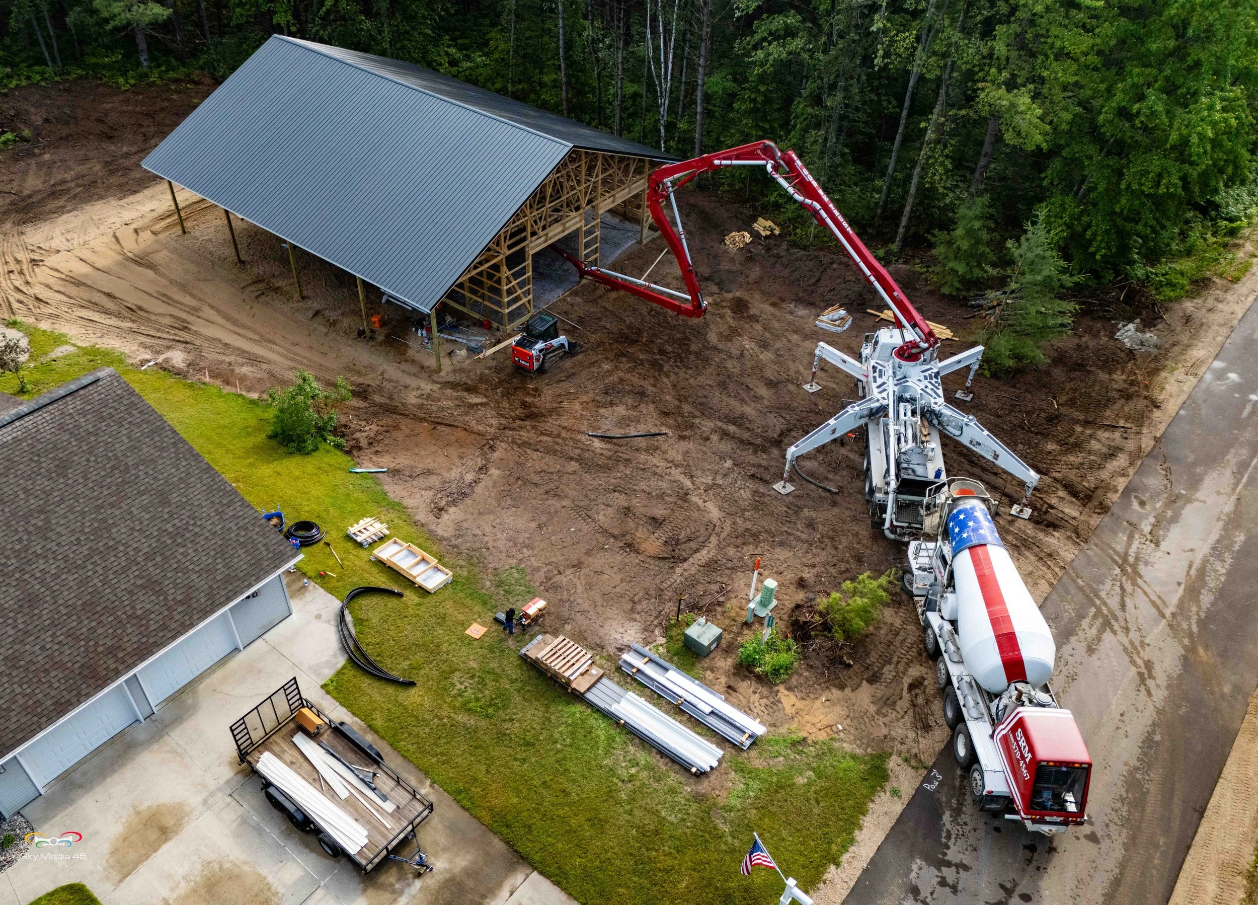 A construction site with a steel building framework under a metal roof, surrounded by trees. A concrete mixer truck is pouring concrete into the building, with construction materials and equipment nearby.