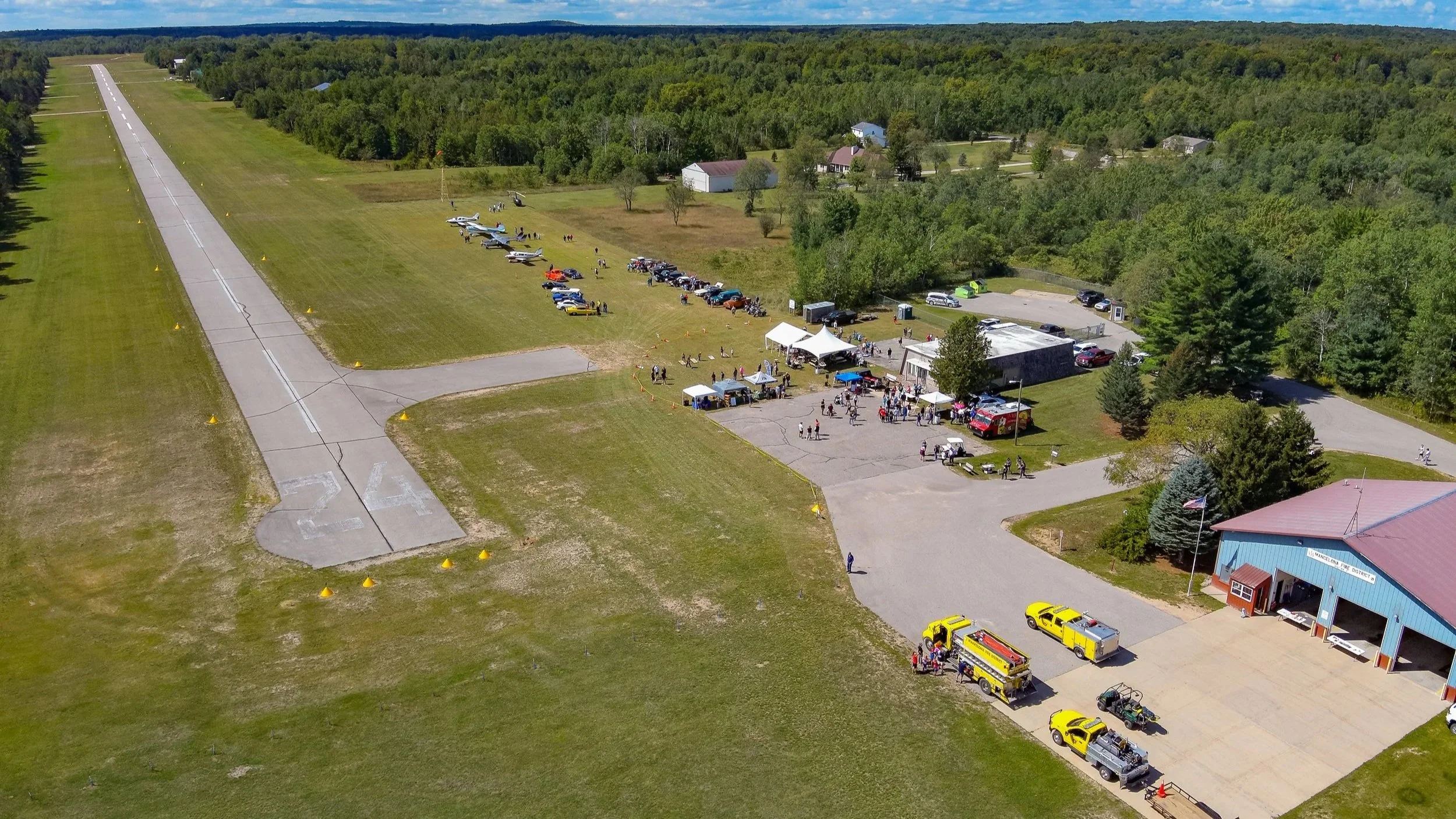 An aerial view of a small airport with a runway and parked small airplanes at Lakes of the North. There is a gathering of people, tents, and emergency vehicles near a building at the edge of the airport, surrounded by green trees and rural landscape.