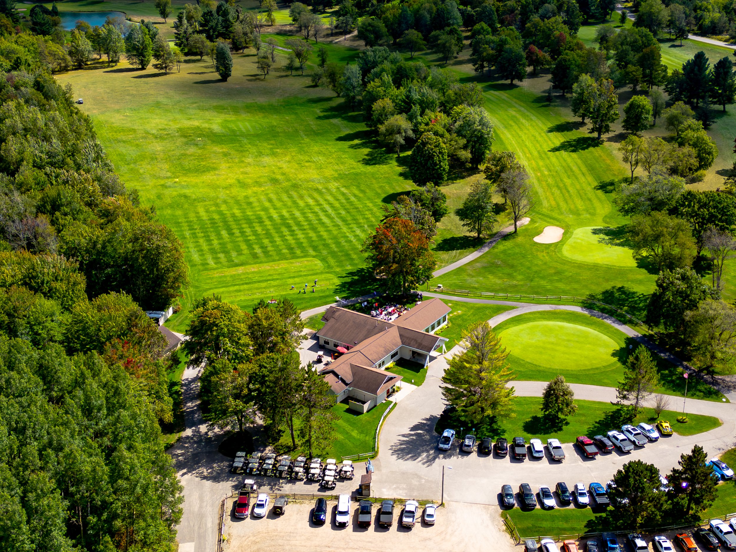 An aerial view of a golf course surrounded by lush green trees, a small pond, and a parking lot filled with cars near a clubhouse.