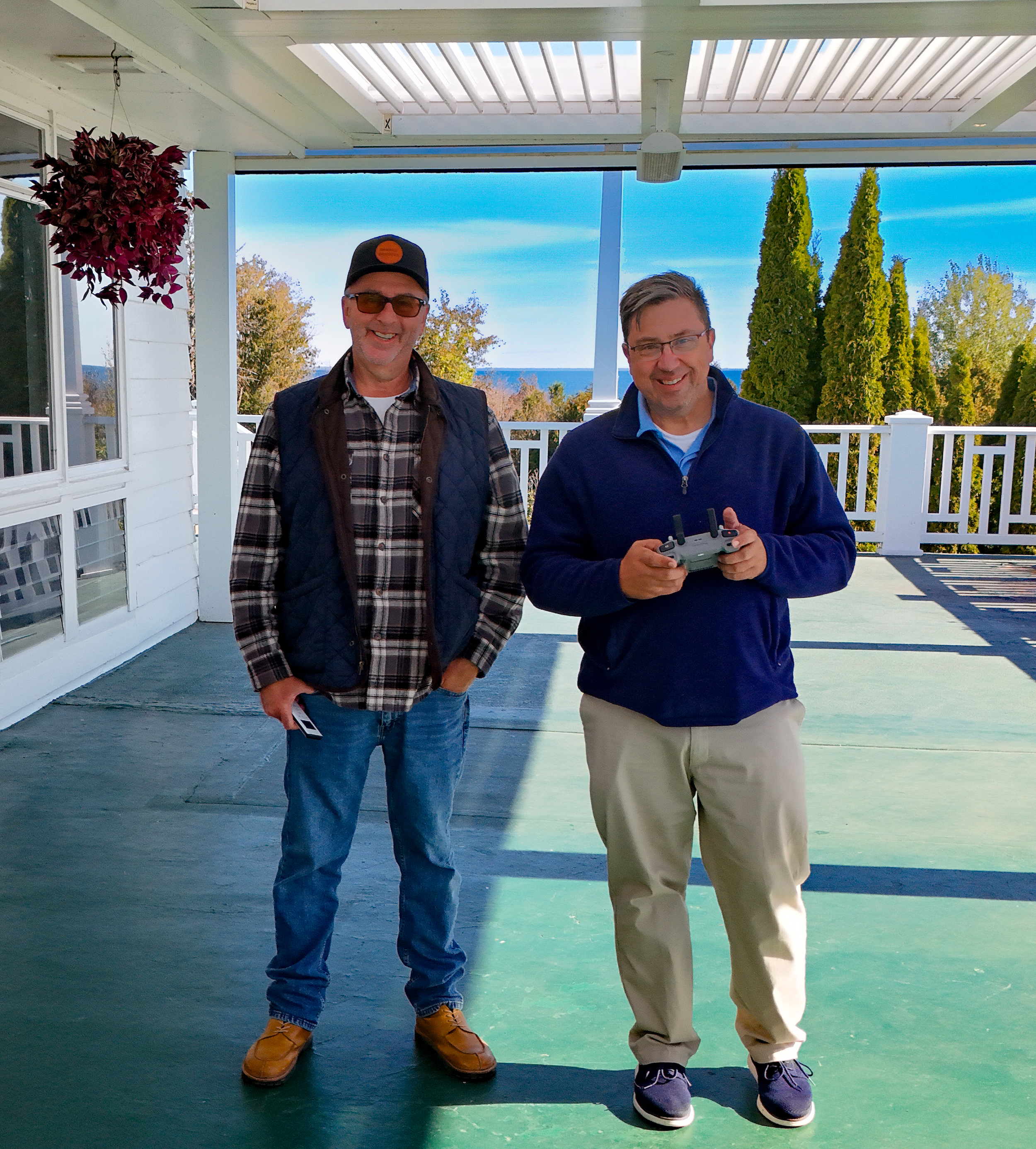 Two men standing on a porch with a view of trees and a blue sky behind them. The man on the left wears glasses, a baseball cap, a plaid shirt, and a dark vest, holding a remote control. The man on the right wears glasses, a blue jacket, and khakis, holding a drone remote control. Both are smiling.